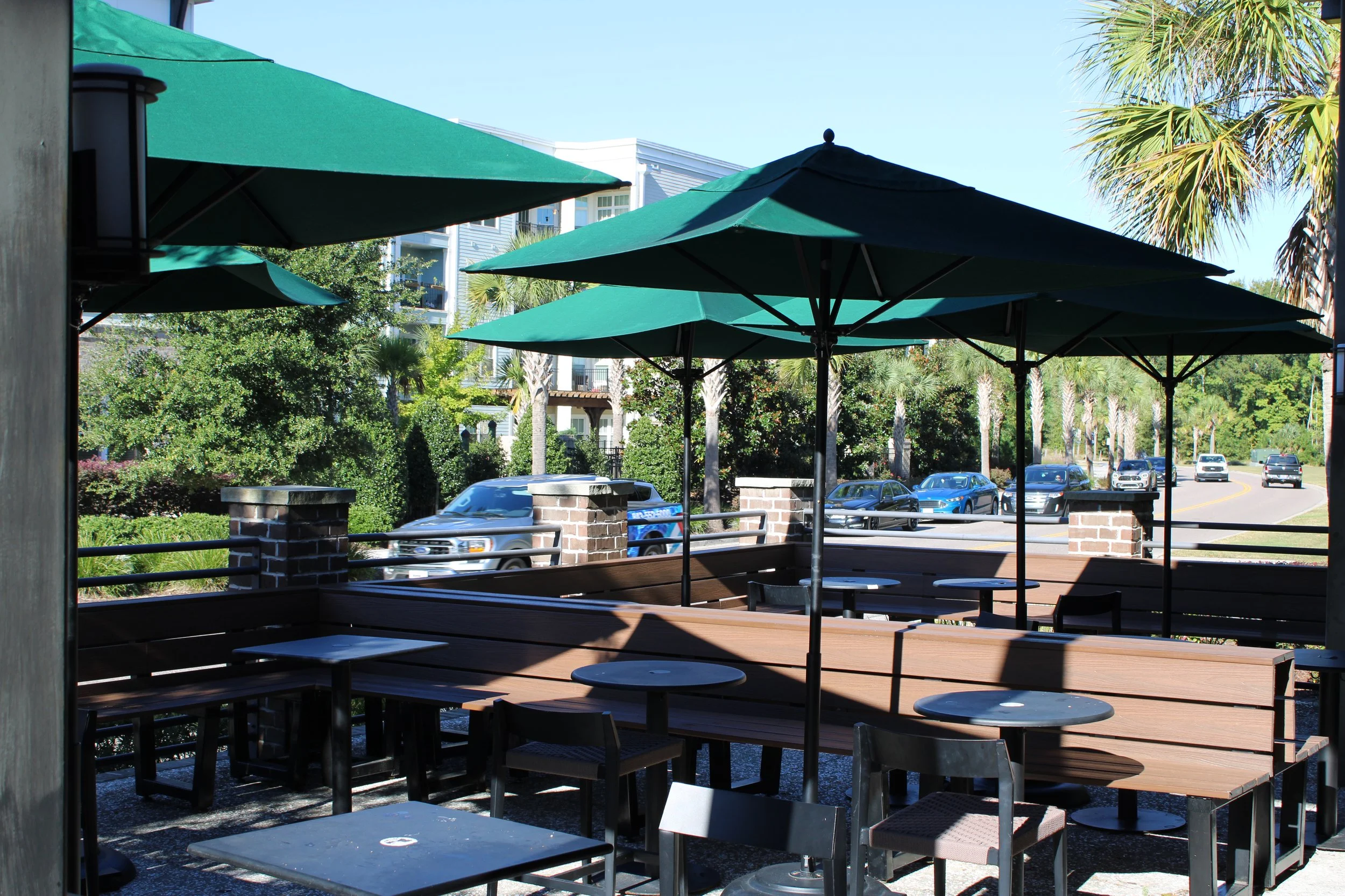 Outdoor patio with green umbrellas, wooden tables and chairs, palm trees, and a parking lot with cars in the background.