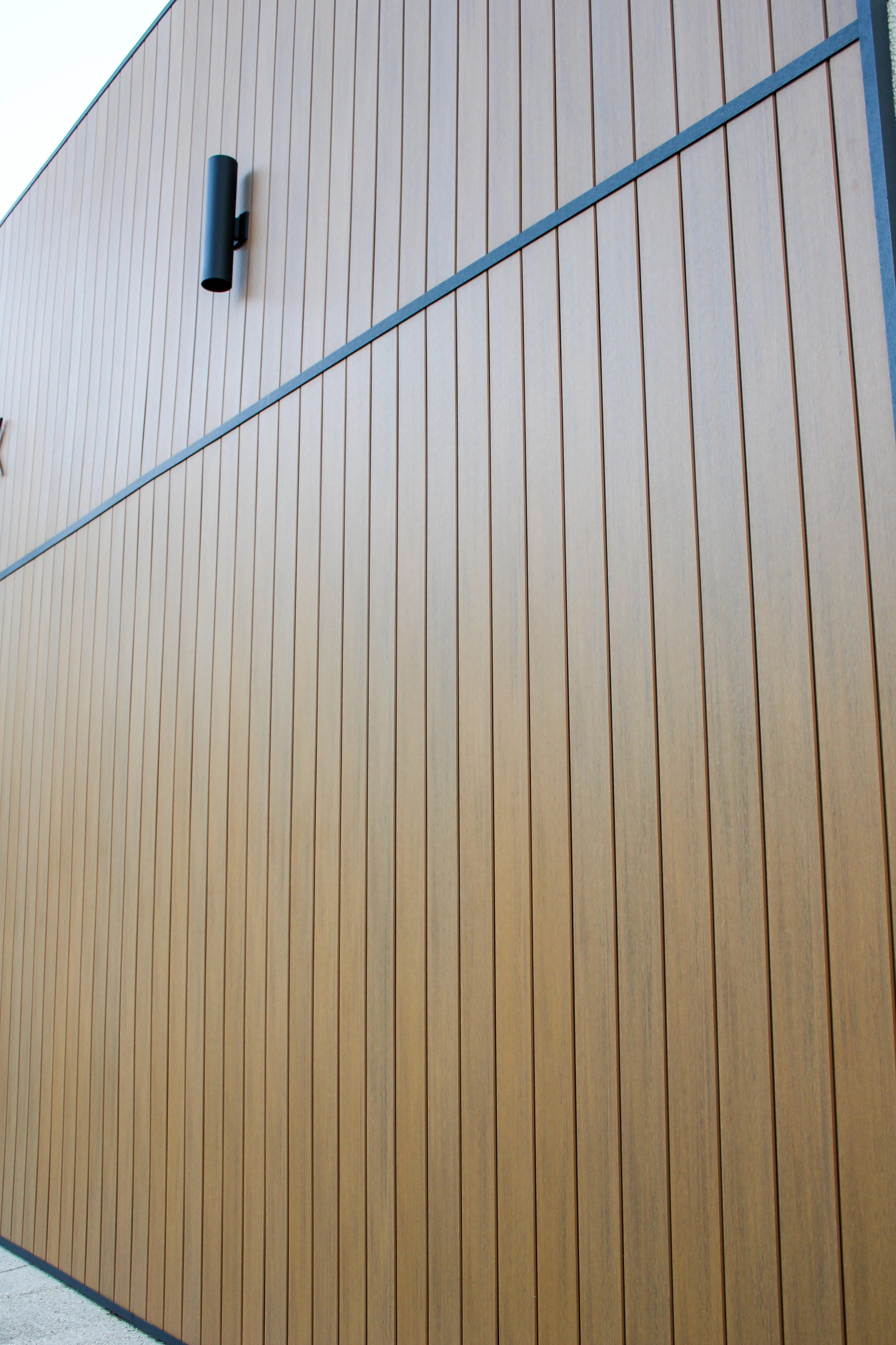 Close-up of a brown wooden exterior wall with vertical panels and a black cylindrical outdoor light fixture mounted on it.