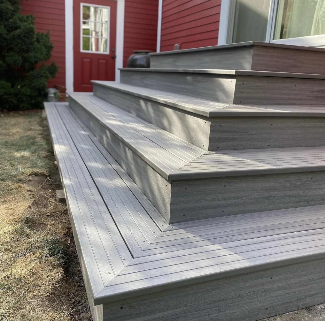 Wooden outdoor steps leading up to a red door with a window, beside a glass window, on a house with red siding. The steps are gray, with a metal strip edge, and the ground around is partially grassy and dirt.