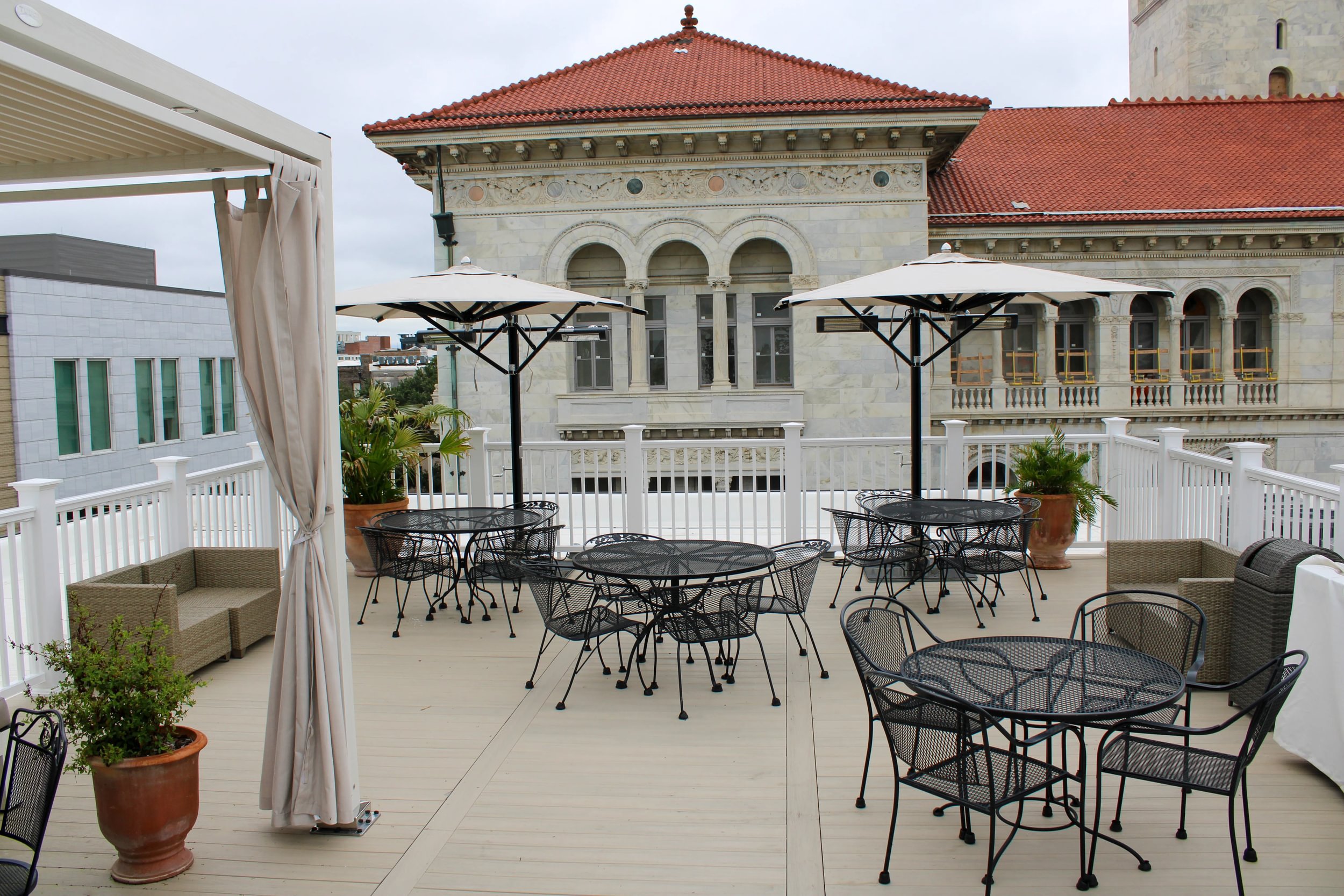 A rooftop terrace with black metal chairs and tables, large potted plants, and white umbrella shades, overlooking historical and modern buildings under an overcast sky.