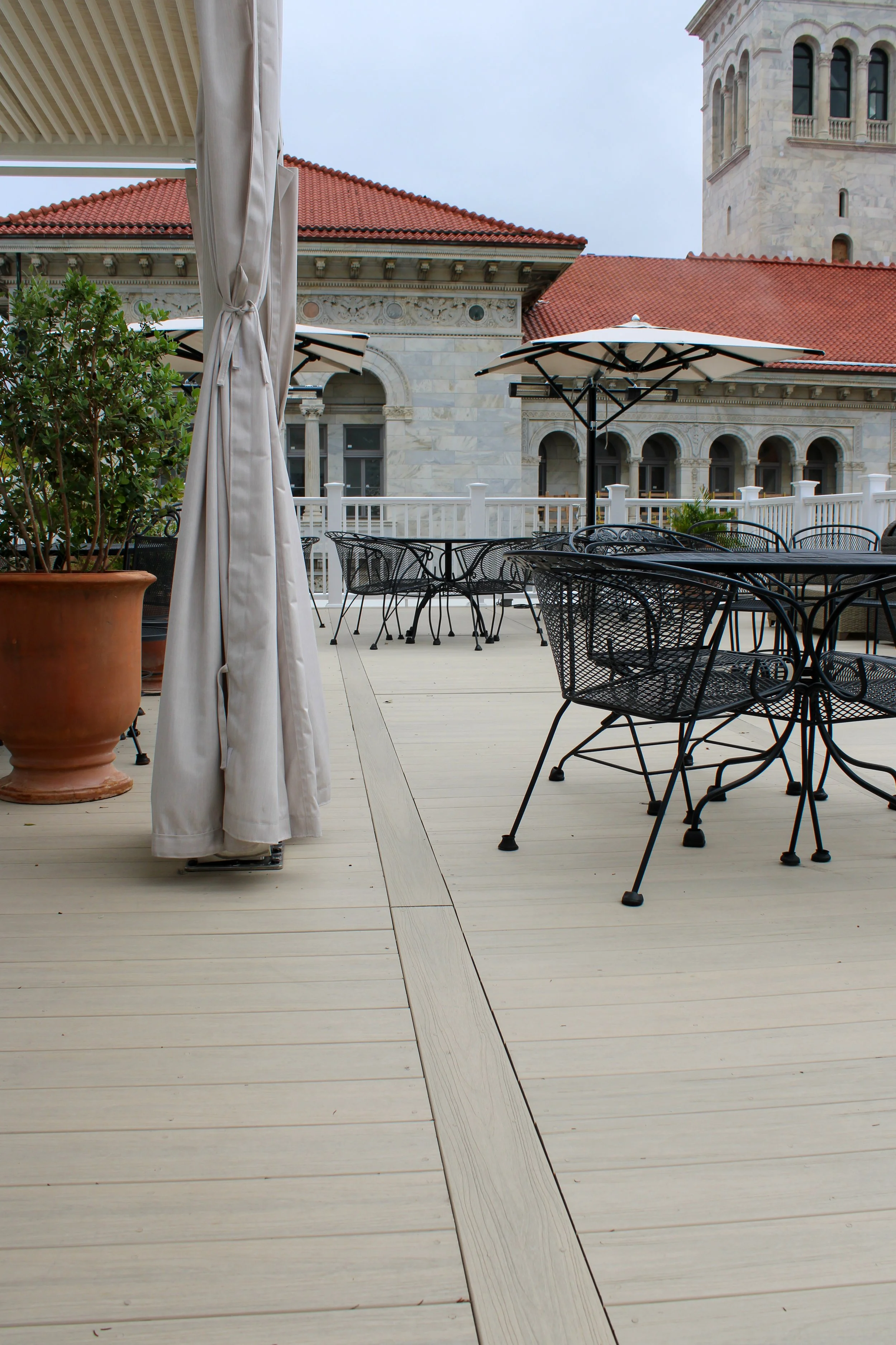 Outdoor patio area with black metal chairs, patio umbrellas, potted plant, and a historic building with red roof tiles and arches in the background.