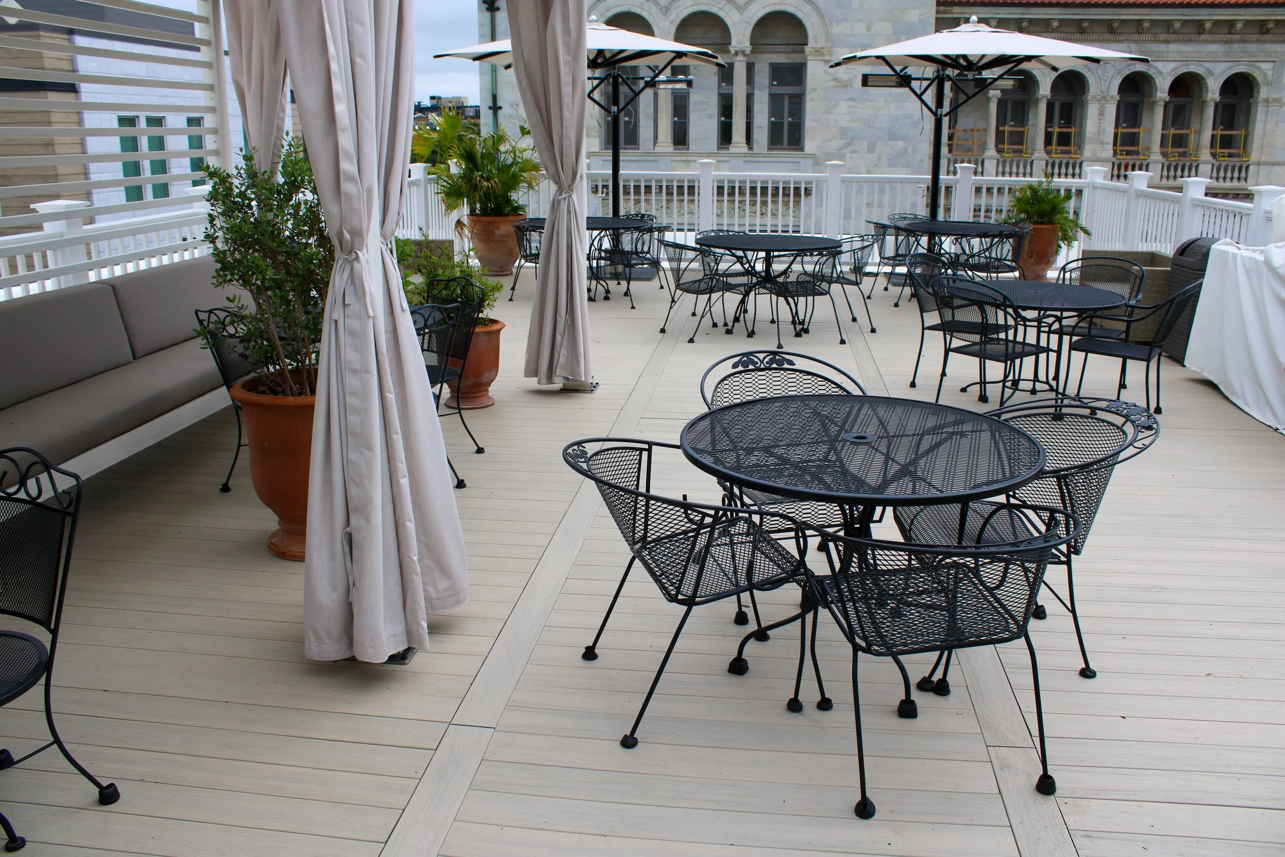 Empty outdoor rooftop patio with black metal tables and chairs, large potted plants, and white curtains tied back. Buildings are visible in the background.