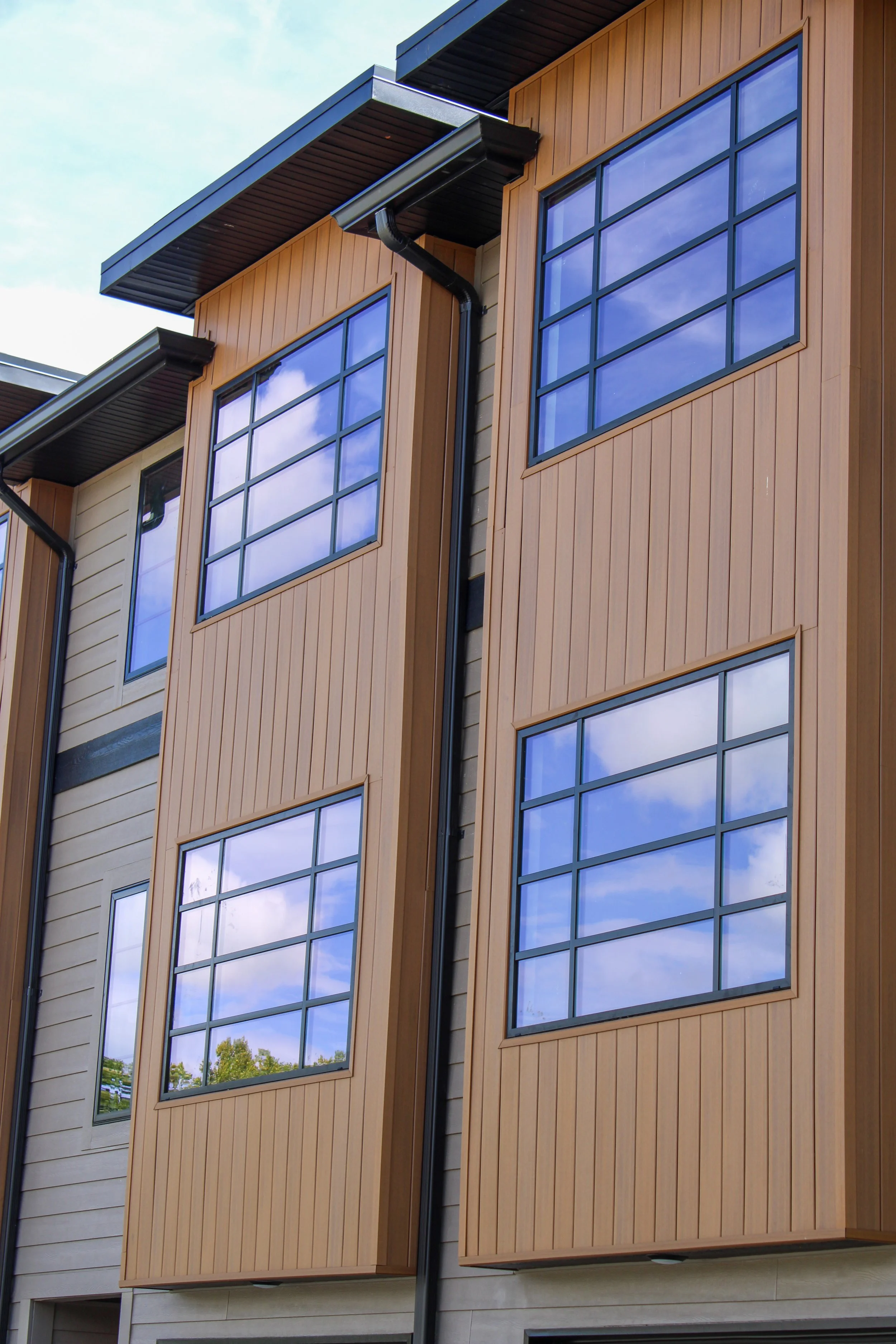 Close-up view of a multi-story modern residential building with large reflective windows and wooden exterior panels, under a blue sky with scattered clouds.