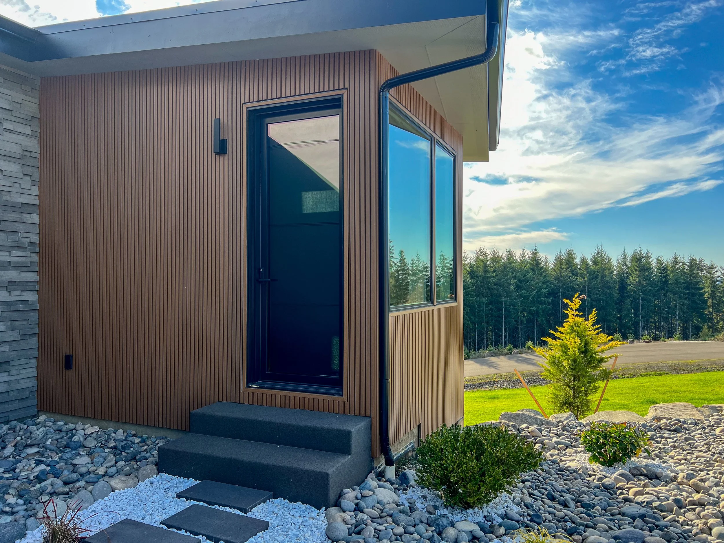 Modern house exterior with wood siding and large glass window, surrounded by landscaped yard with rocks, small tree, and open green space under a partly cloudy sky.