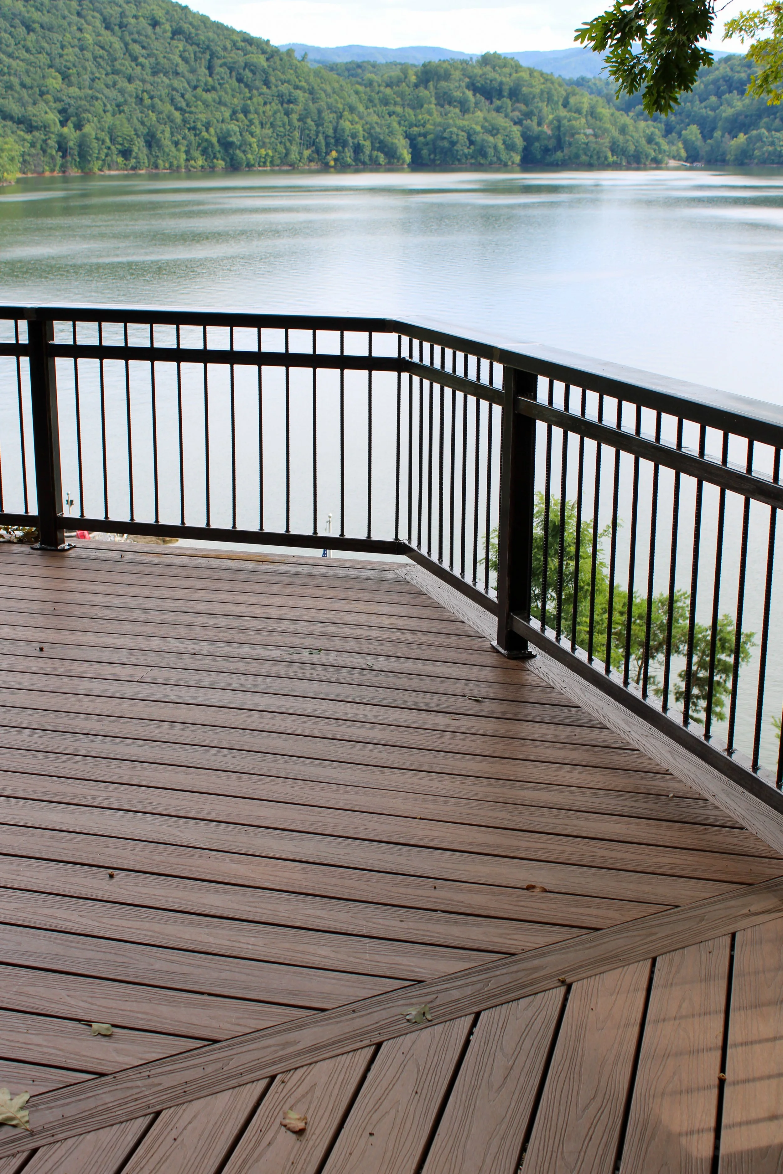 View from a wooden deck with a black metal railing overlooking a lake, surrounded by green mountains and trees, with cloudy sky