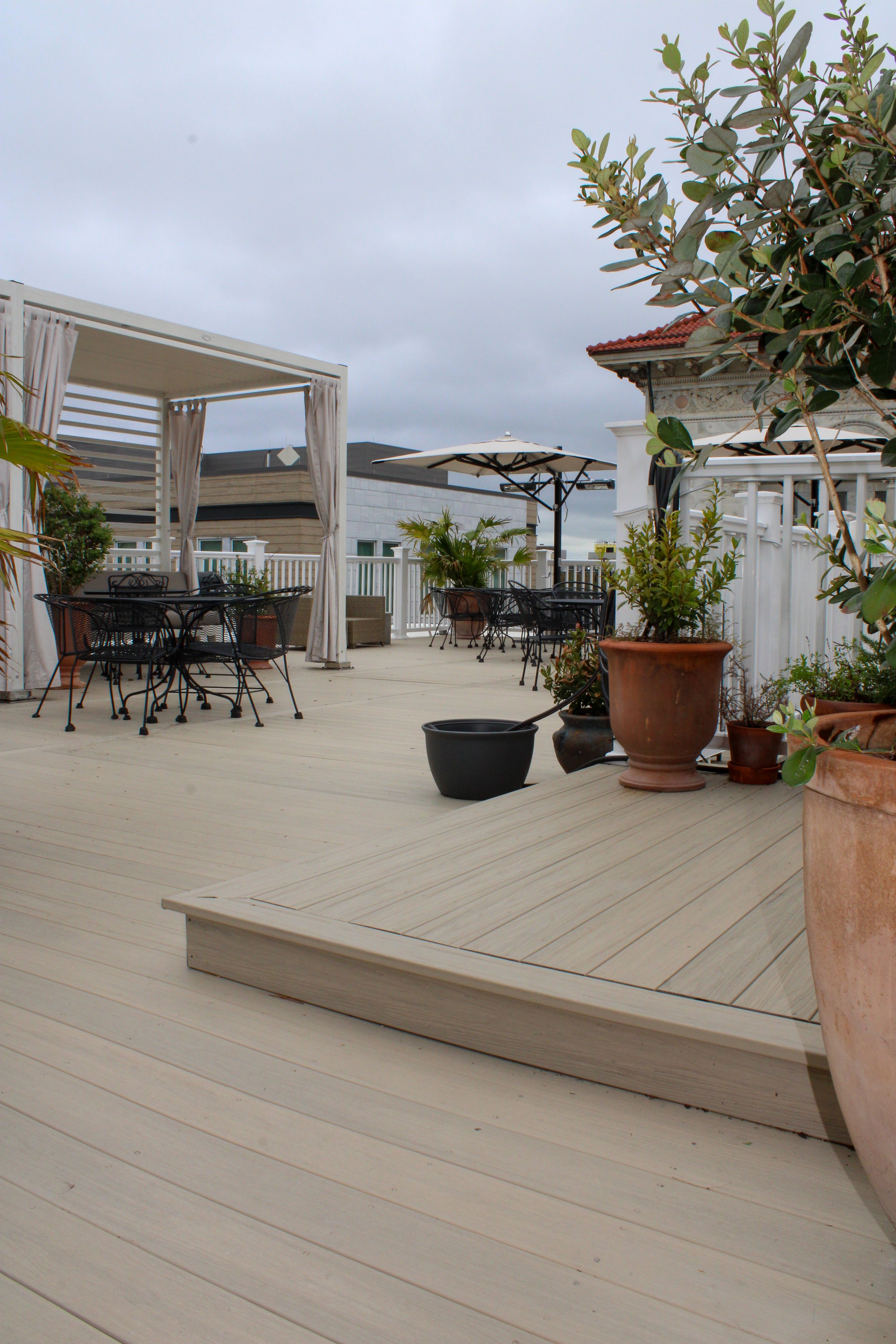 An outdoor patio area with wooden flooring, potted plants, black metal tables and chairs, a white pergola with curtains, a large umbrella, and a white railing fence.