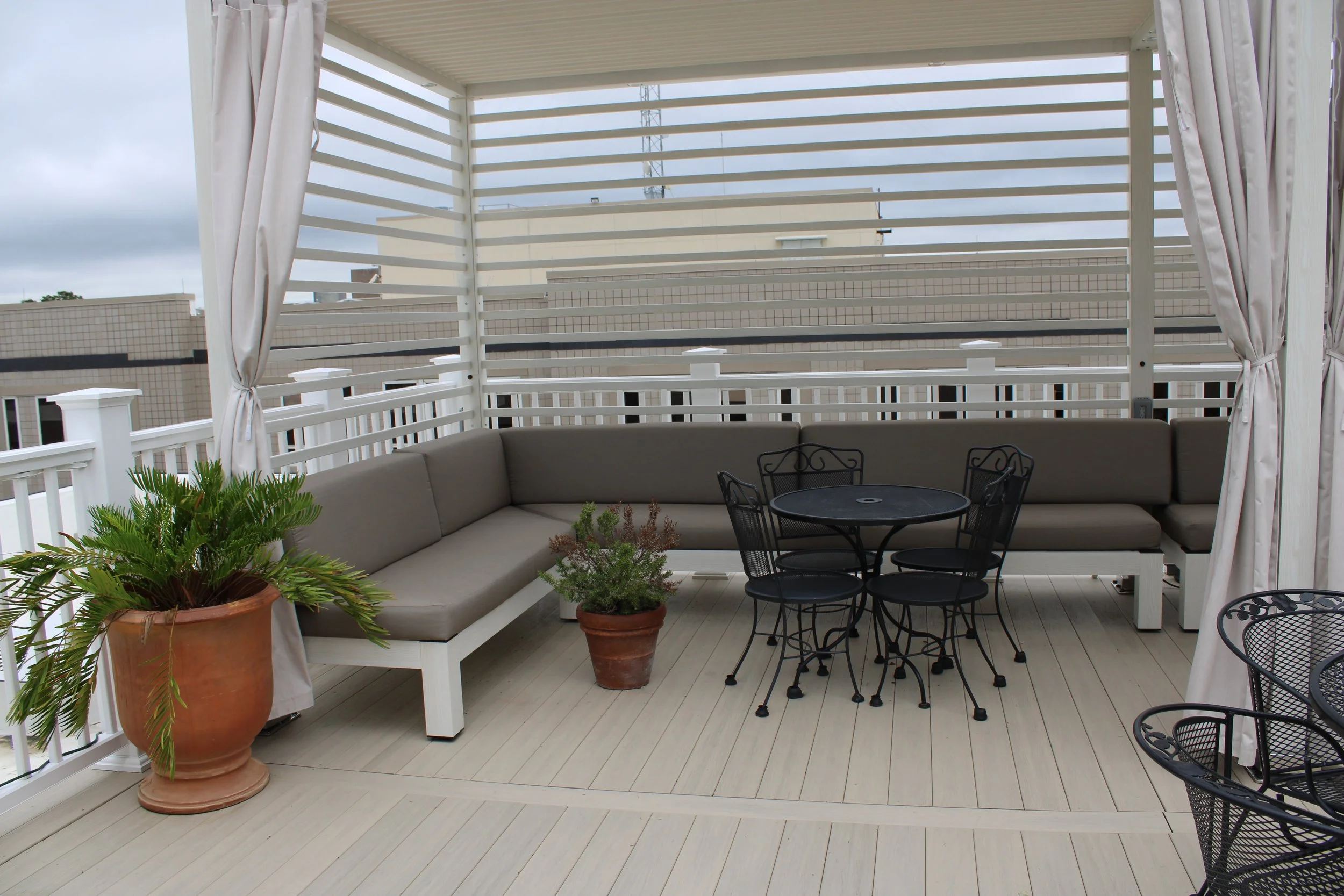A covered outdoor balcony with a beige sectional sofa, a small black metal table with four matching chairs, and several potted plants including a large terracotta pot with green foliage. White curtains frame the space, and white striped privacy fenci
