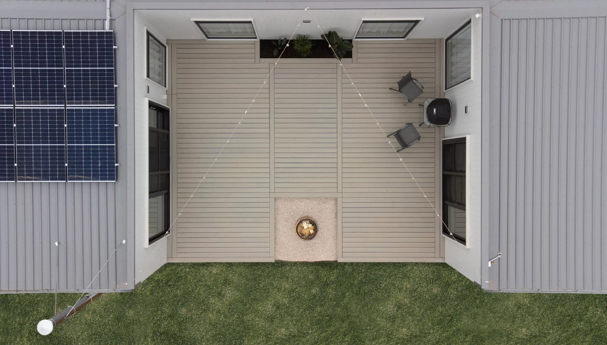 Top-down view of a house's backyard patio with beige wood decking, framed by house walls with solar panels on the left, and a small fire pit in the center. There are two chairs, a table, and a few potted plants in the patio area.