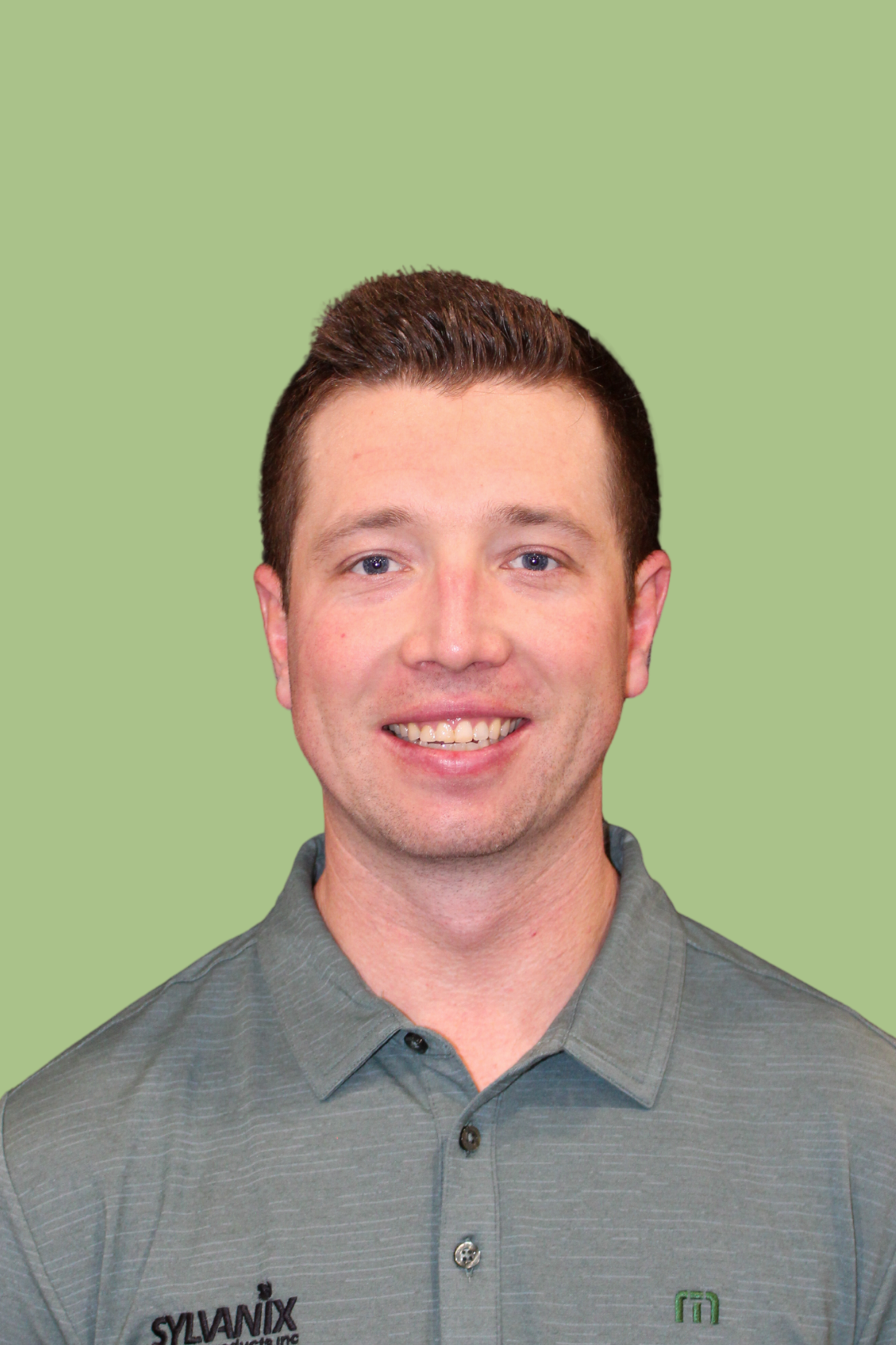 A young man with short brown hair, blue eyes, and a light complexion, wearing a gray collared shirt with the logo and text 'SYLVANIX' on the chest and a green emblem, smiling against a solid light green background.