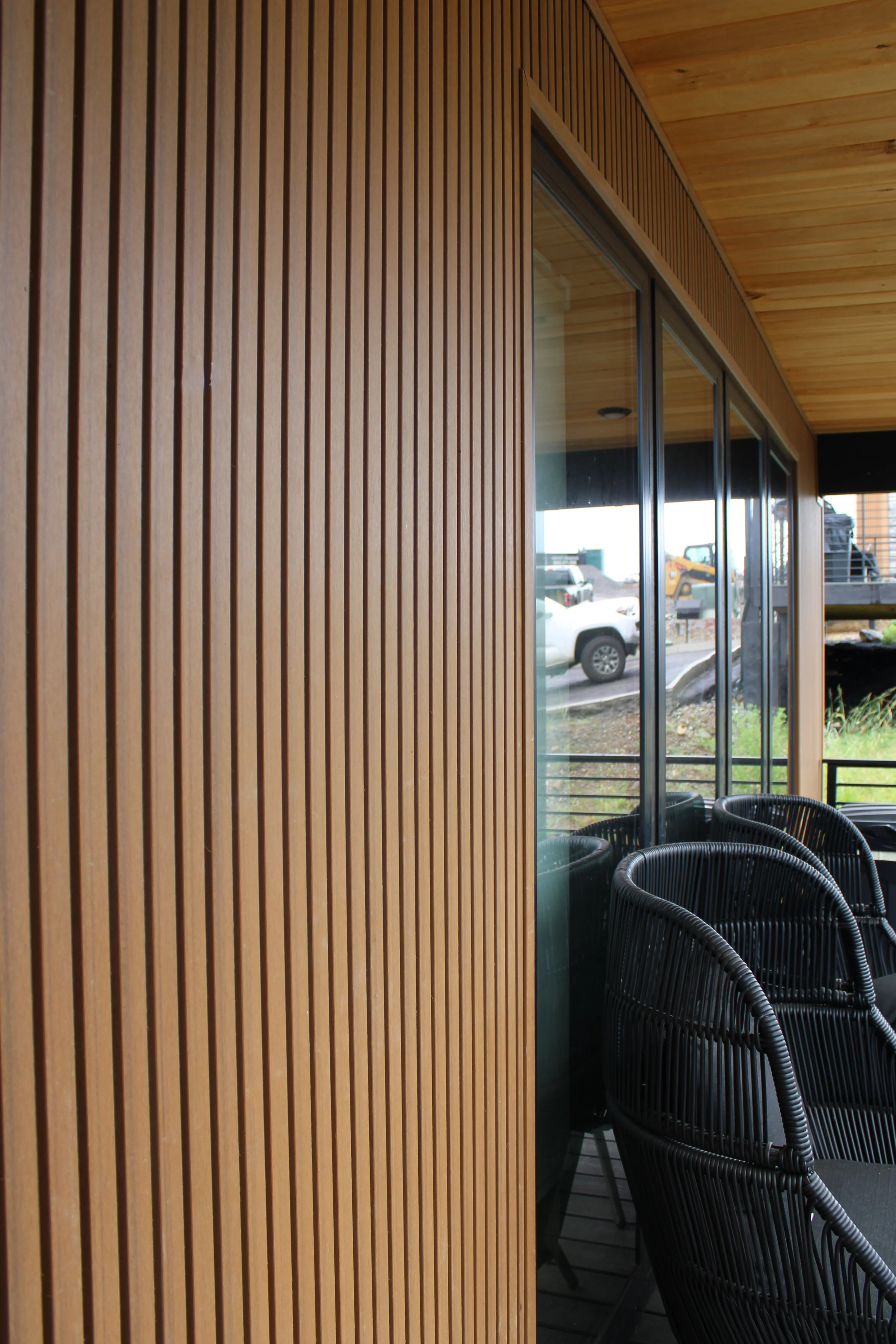Wooden wall and ceiling of an outdoor patio area with glass sliding doors and black outdoor chairs. Cars are visible outside.