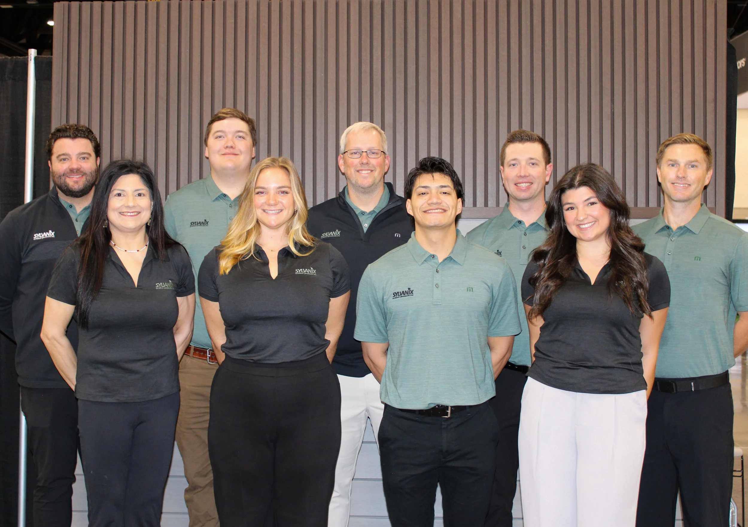 Group photo of nine smiling people, six men and three women, posing indoors against a wooden paneled wall. They are wearing business casual clothes, with some in black and others in green or gray shirts, and some have logos on their shirts.