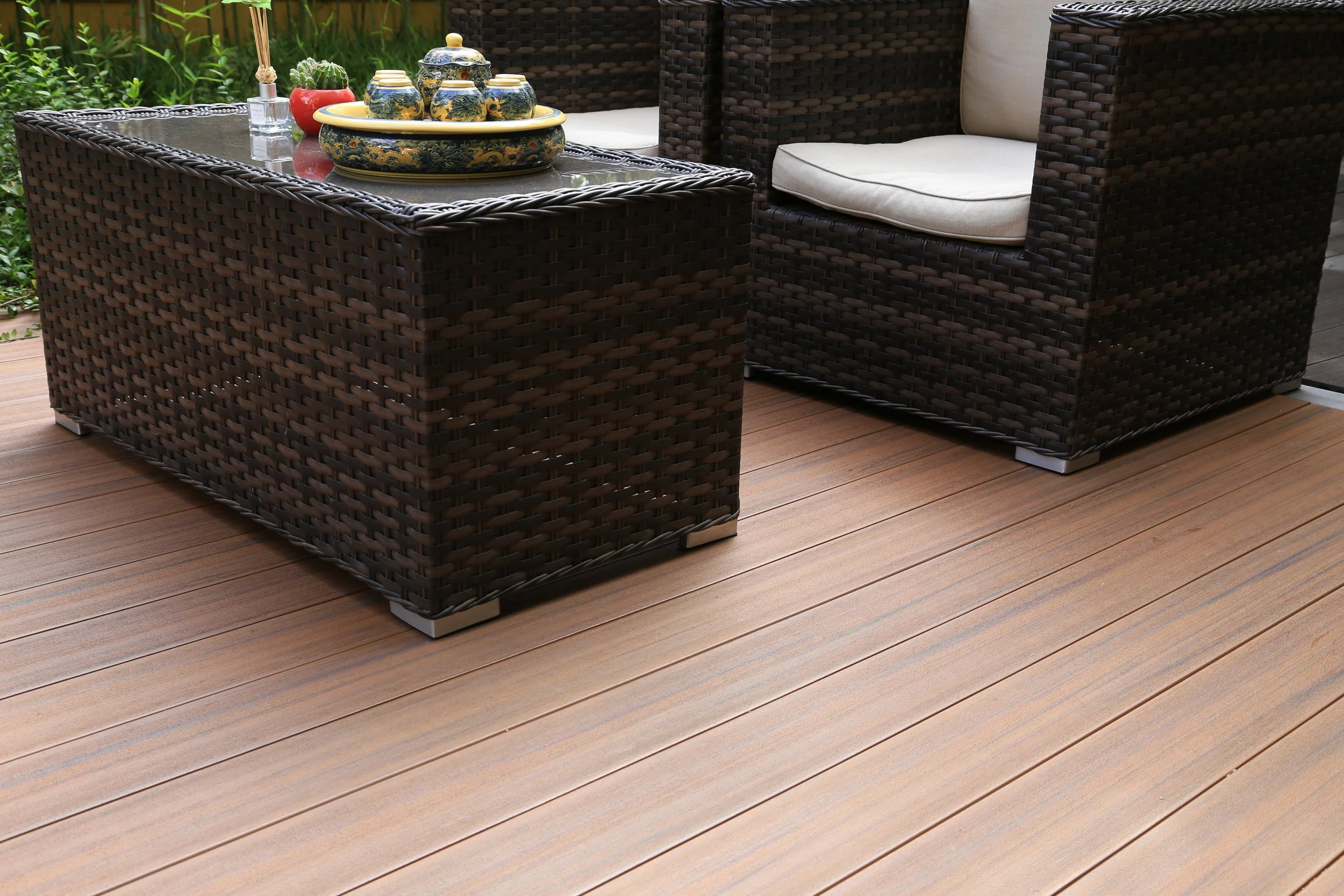Outdoor patio furniture set with a rectangular wicker table and two matching wicker armchairs with cushions, placed on a wooden deck near a garden with green plants.