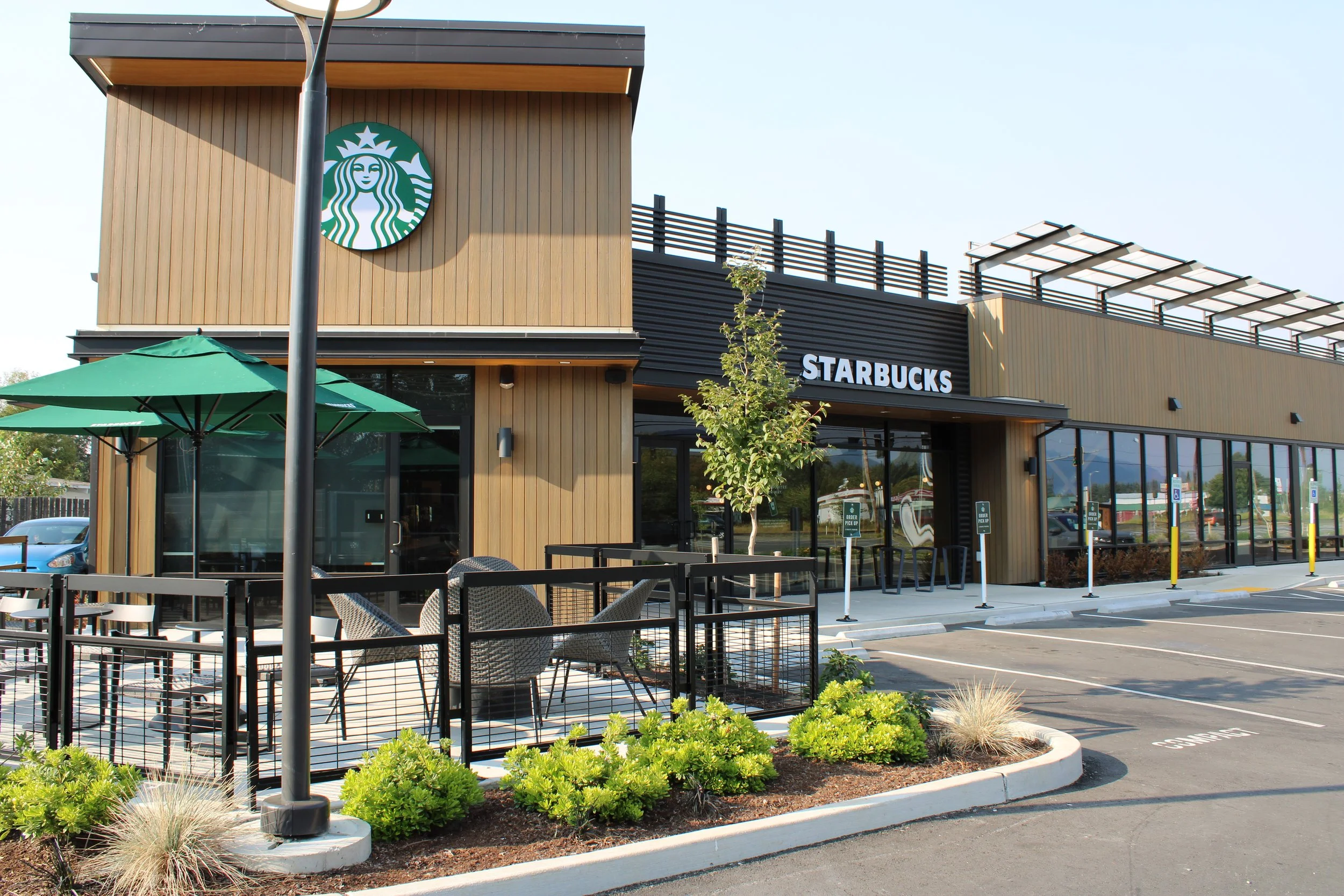 Exterior of a Starbucks coffee shop with outdoor seating, umbrellas, and landscaping in a parking lot.
