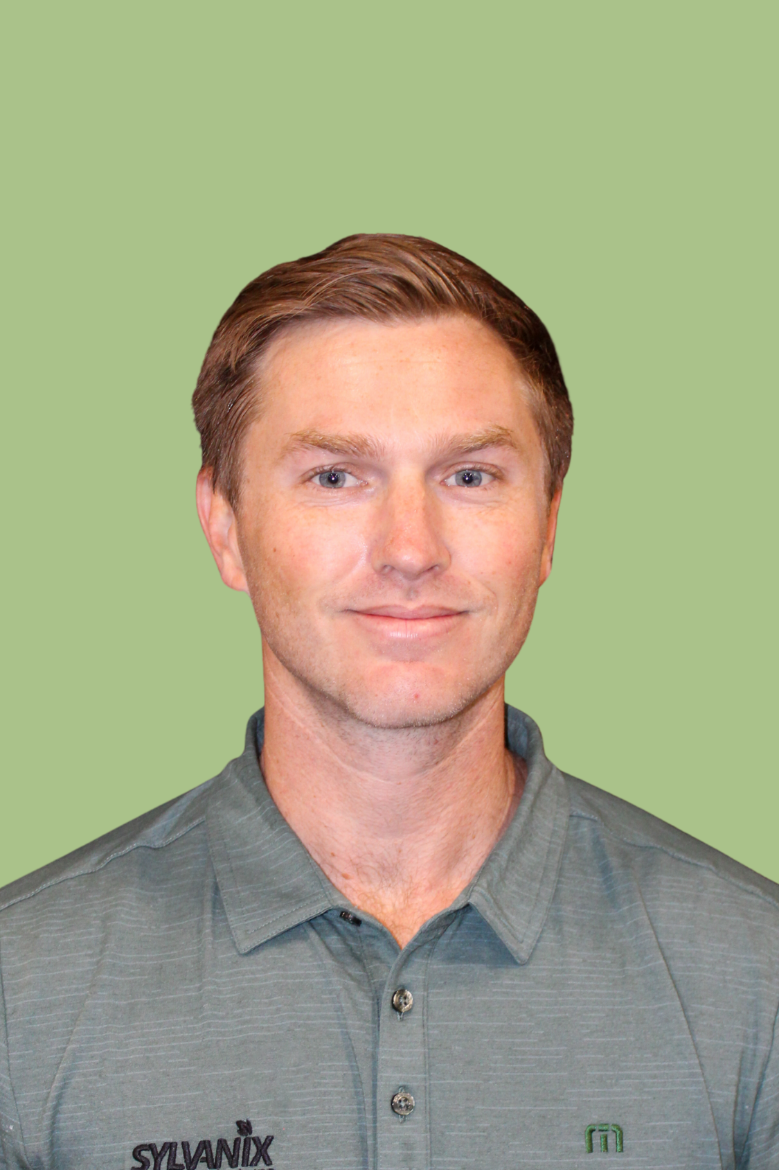 A man with short brown hair and blue eyes, wearing a gray collared shirt with the logos 'SYLVANIX' and a green emblem, standing in front of a plain green background.