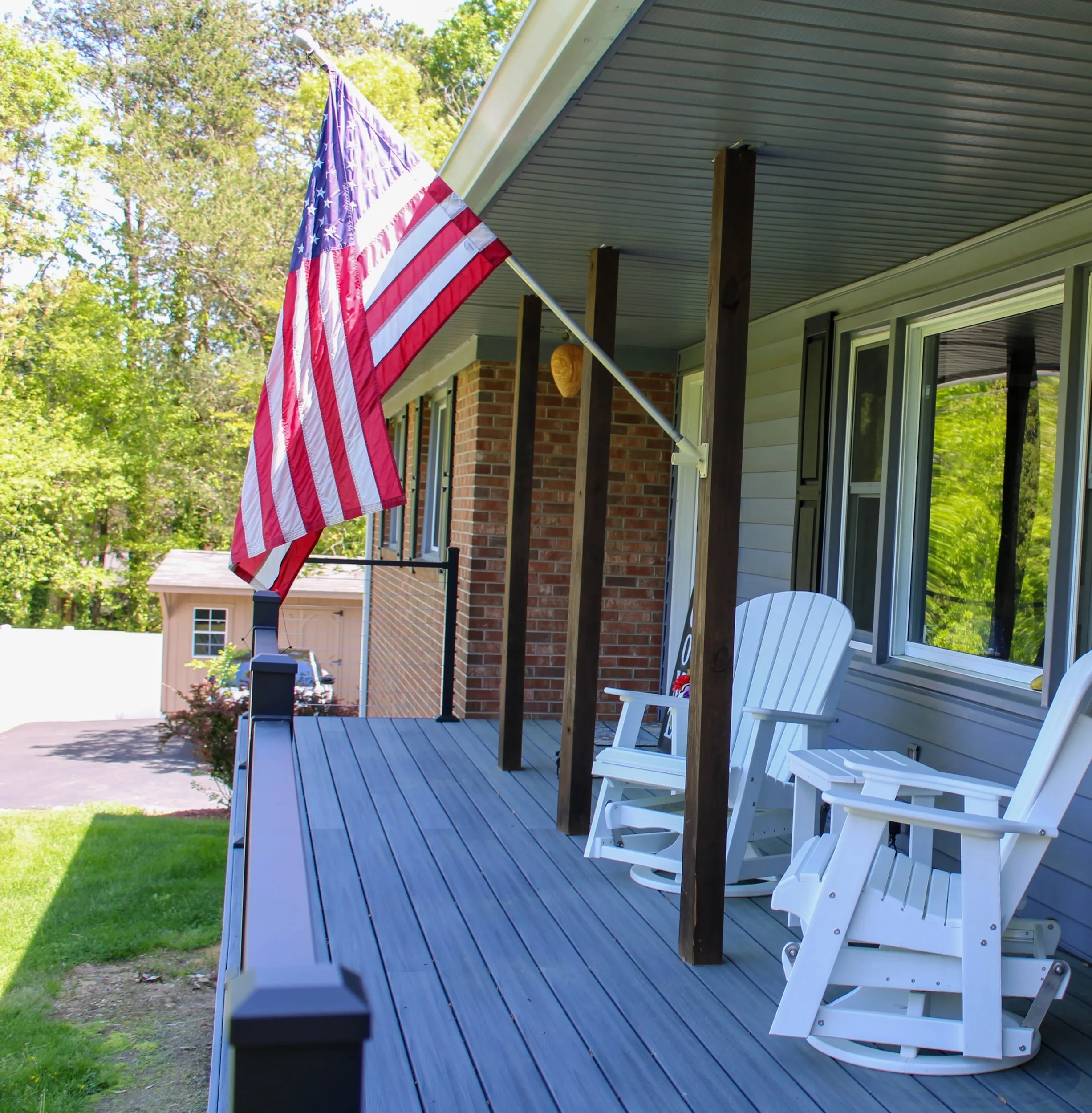 A house porch with blue wooden flooring, white rocking chairs, an American flag hanging from a pole attached to the house, and greenery including trees in the background.
