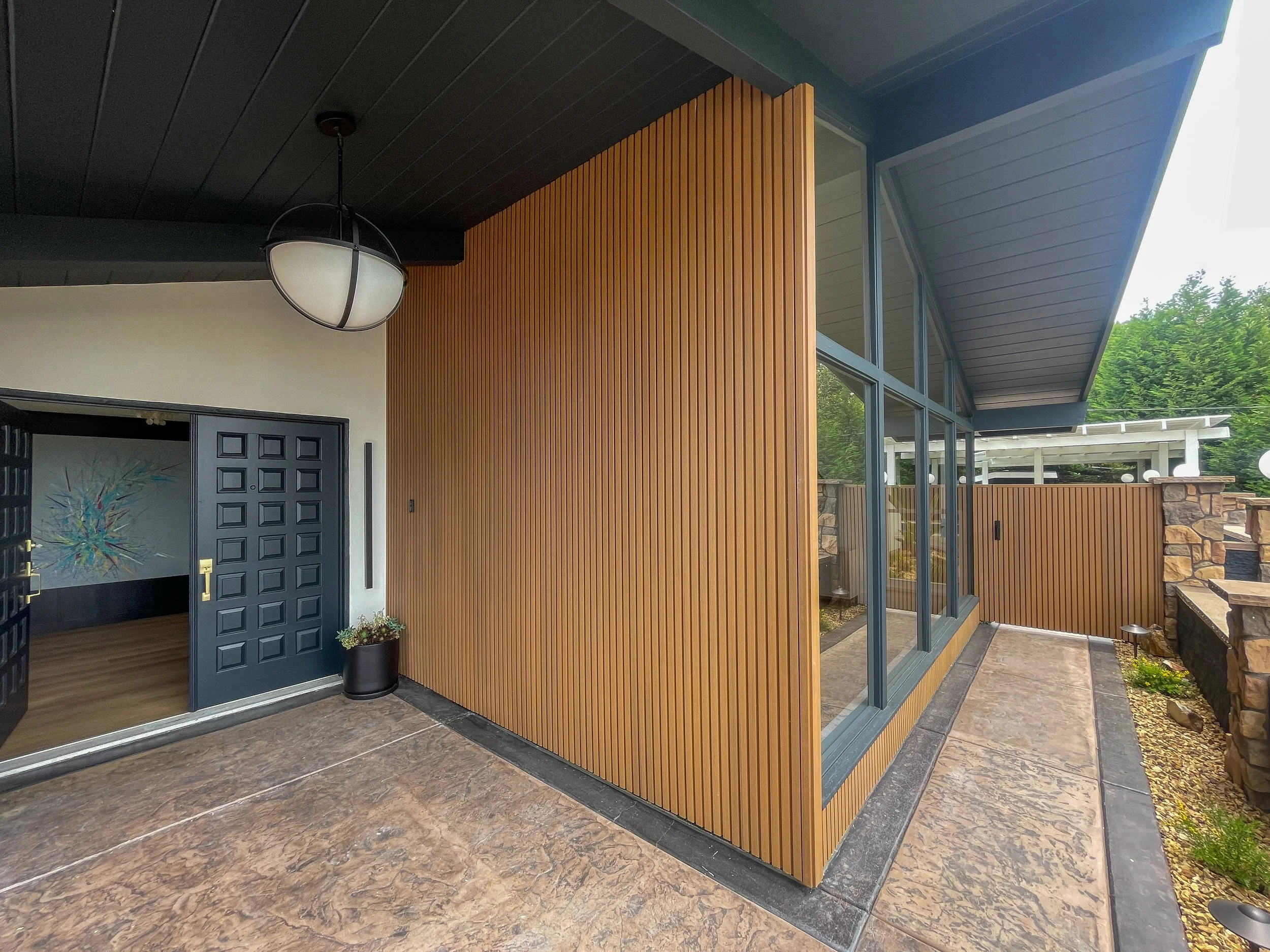 Modern house exterior featuring a wood-paneled wall, large glass windows, and a black front door with geometric design. There is a black potted plant near the door and a textured stone floor, with greenery in the background.