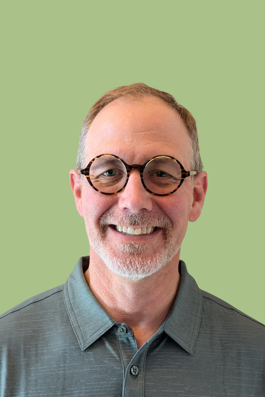 A smiling man with glasses, gray hair, and a beard wearing a gray collared shirt.