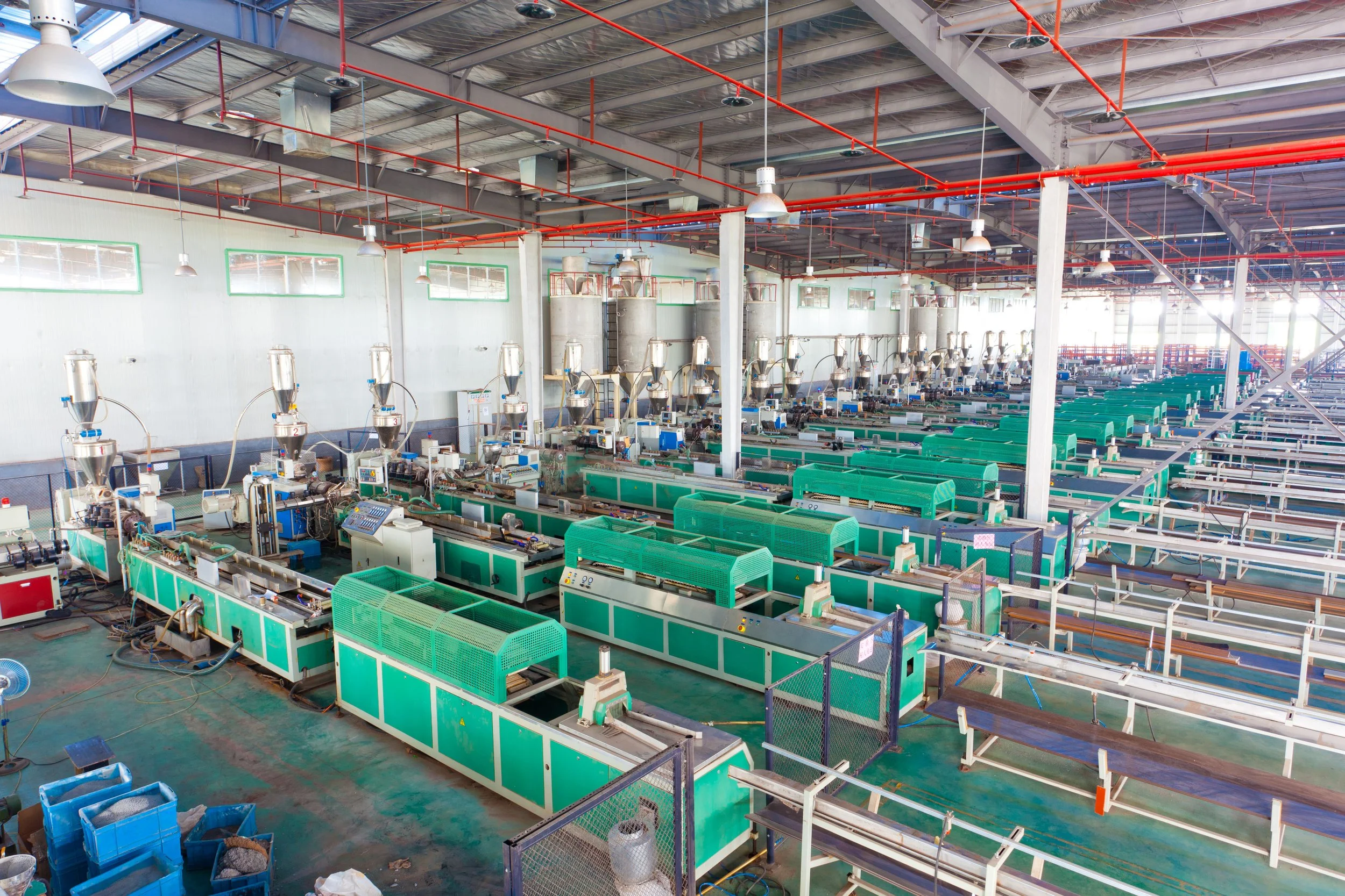 Industrial manufacturing facility with green machines, stainless steel containers, and overhead red piping in a large, well-lit warehouse.