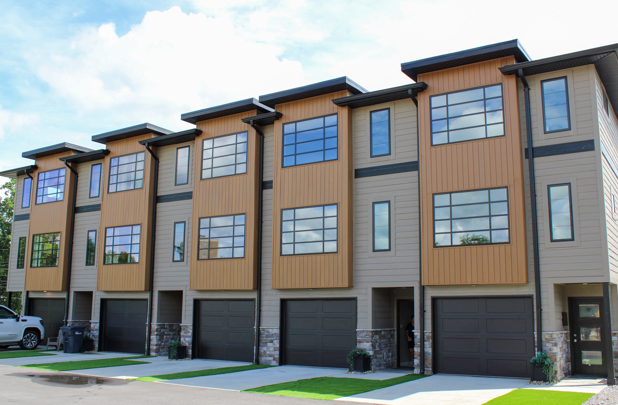 Modern multi-story residential building with large windows, garage doors, and a small lawn in front, under a partly cloudy sky.