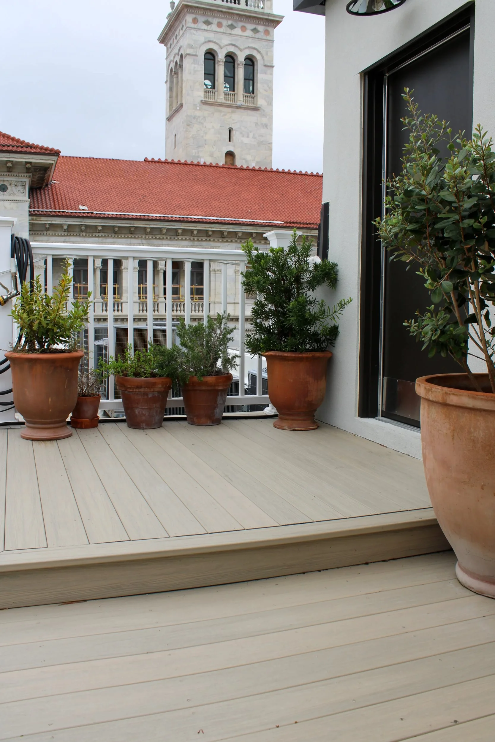A rooftop balcony with several potted plants and a view of a church tower with arches and a red tiled roof.