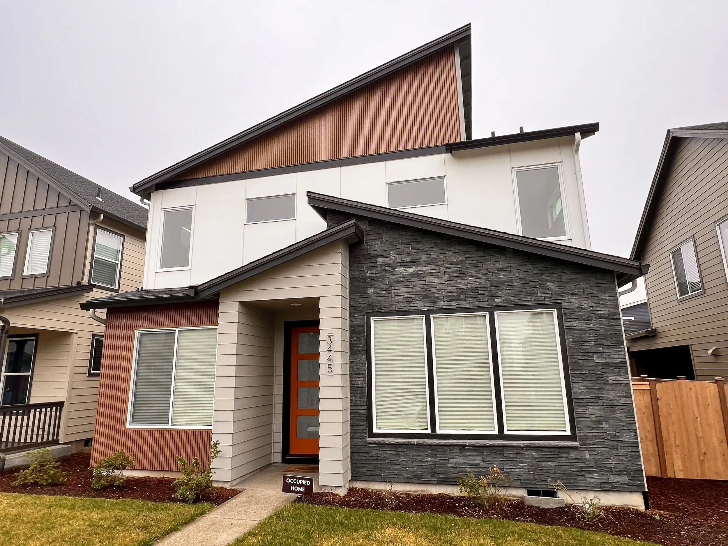 Modern multi-story house with a mix of white, black, and wooden exterior finishes, large windows, and a small front yard with a concrete pathway and a sign that reads 'Occupied Home'.