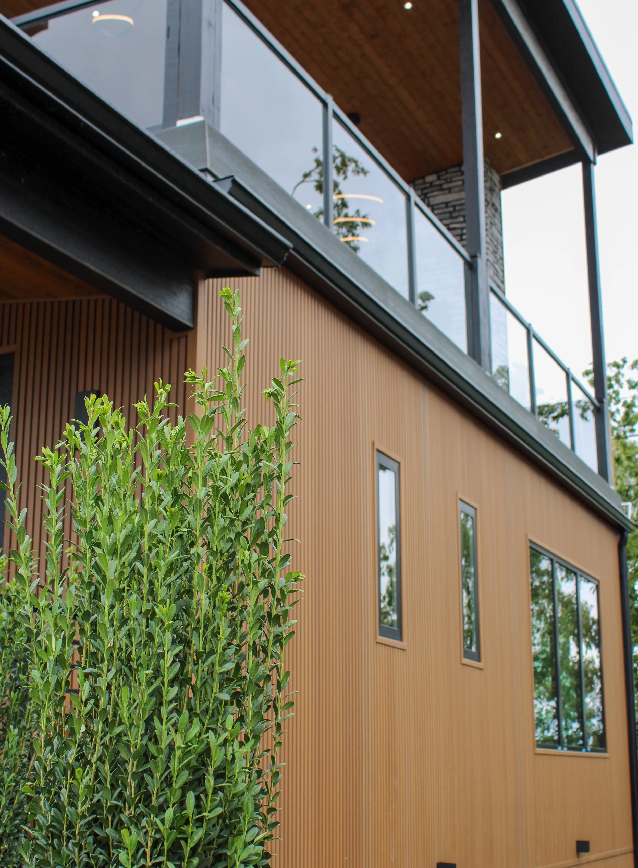 Close-up view of a modern house with a wooden exterior, large windows, a balcony with glass railing, and greenery in the foreground.