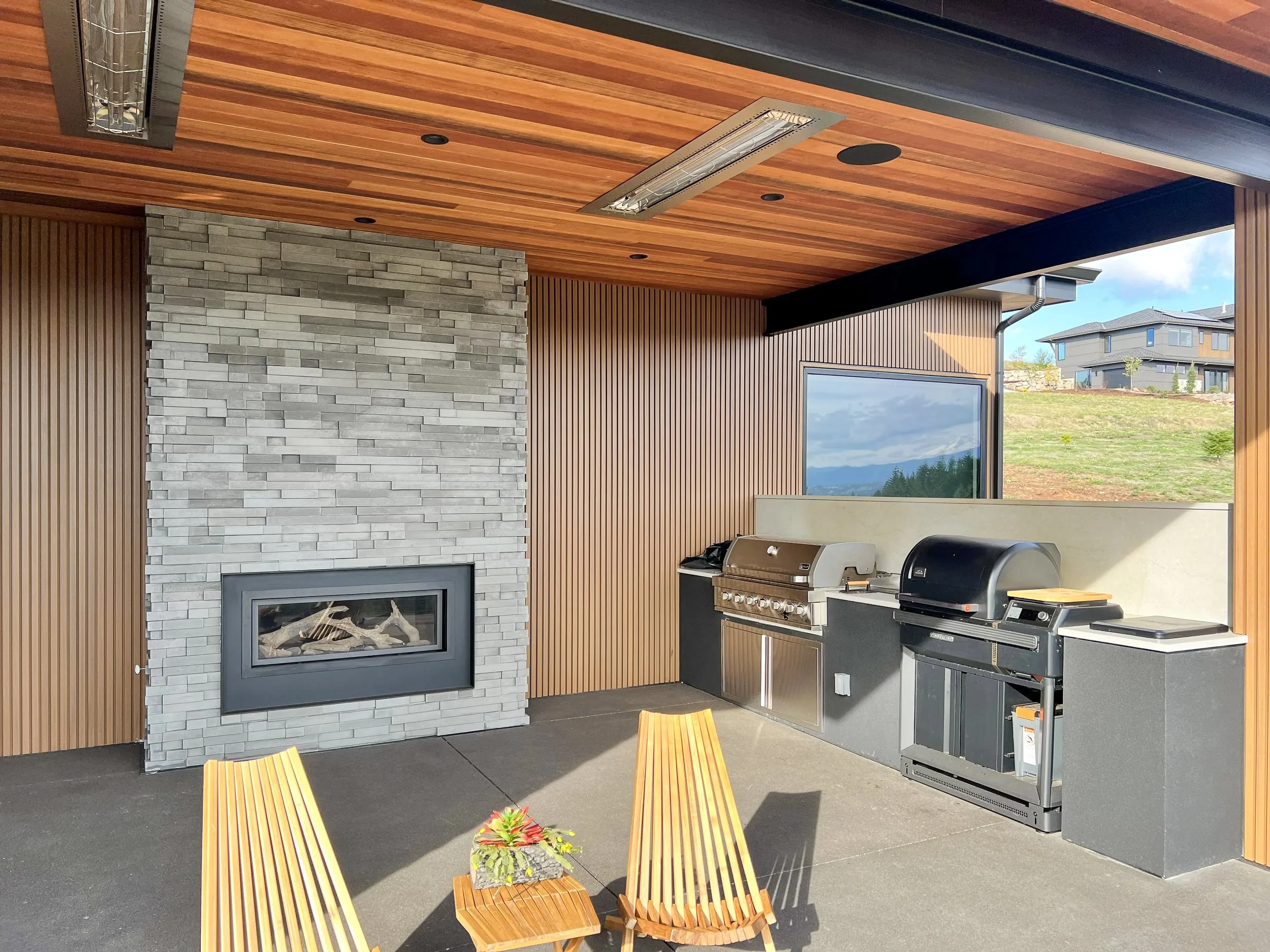 Outdoor kitchen with built-in grill, stone fireplace, and wooden ceiling, with two wooden chairs and a small table with plants.