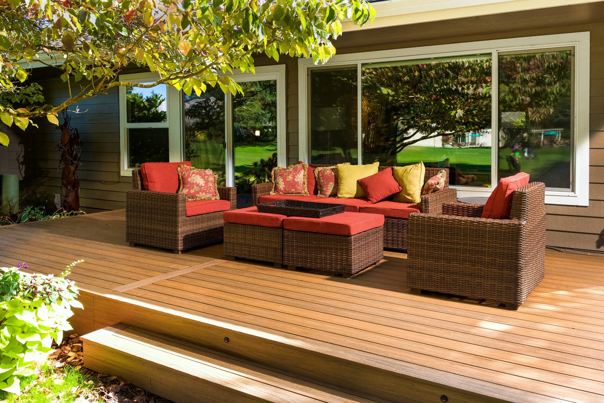 A backyard patio with wooden decking and outdoor wicker furniture that has red and yellow cushions. A tree with green leaves and some flowering plants are visible around the patio, and there is a house in the background with large glass windows reflecting trees and sky.