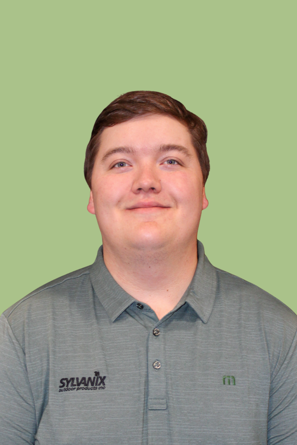 A young man with short brown hair, blue eyes, and fair skin smiling, wearing a gray polo shirt with logos, against a plain green background.