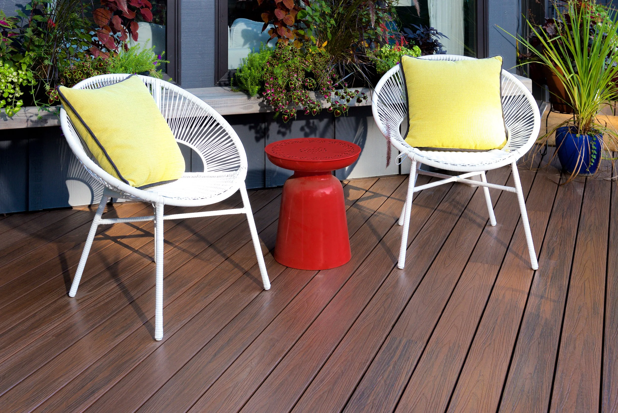 A small outdoor patio with two white circular chairs with yellow cushions, a red small side table, and potted plants on a wooden deck in front of a house with large windows and dark exterior walls.