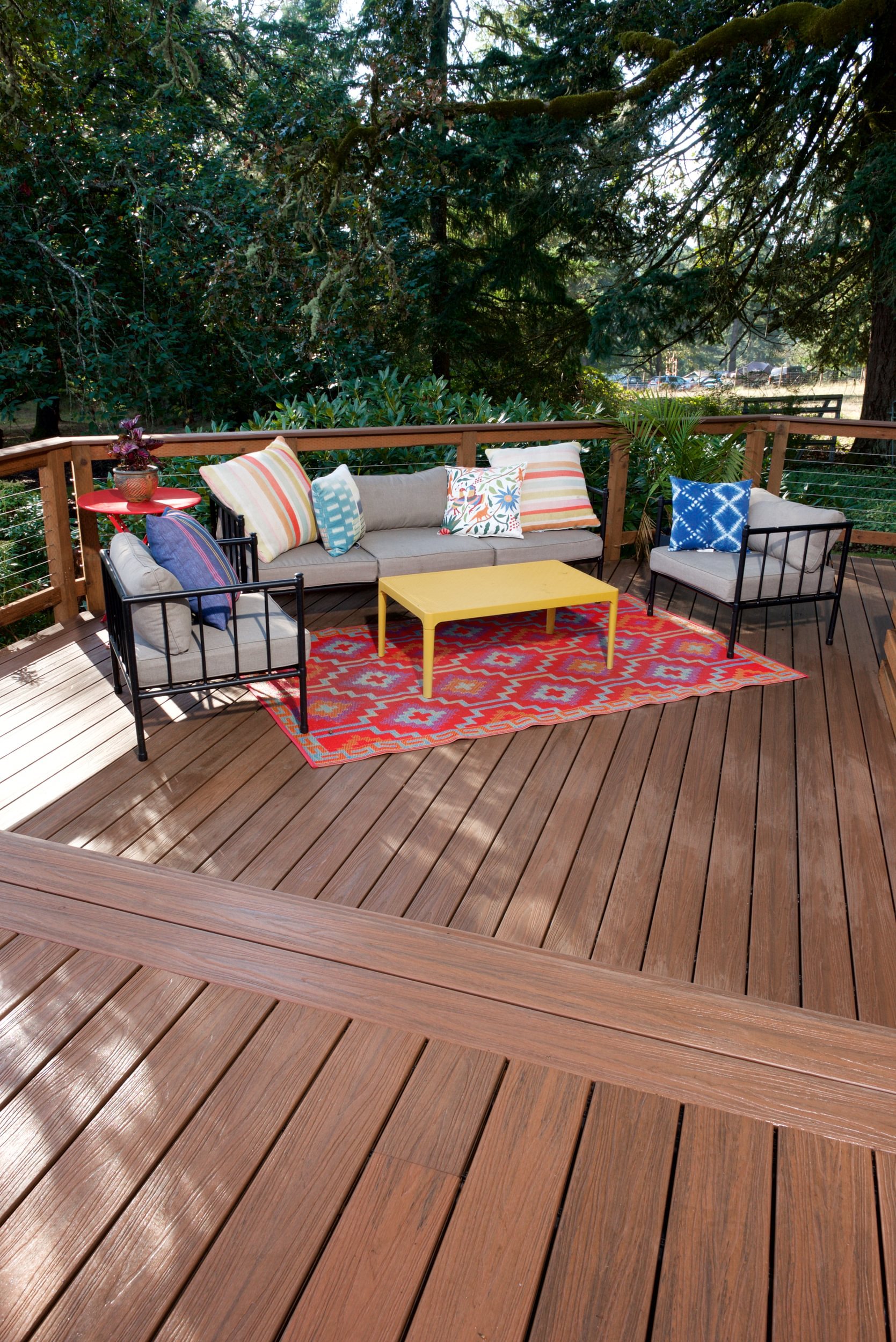 Wooden deck with outdoor seating area including a beige sofa, two black metal chairs, a yellow coffee table, and colorful pillows, surrounded by lush green trees.