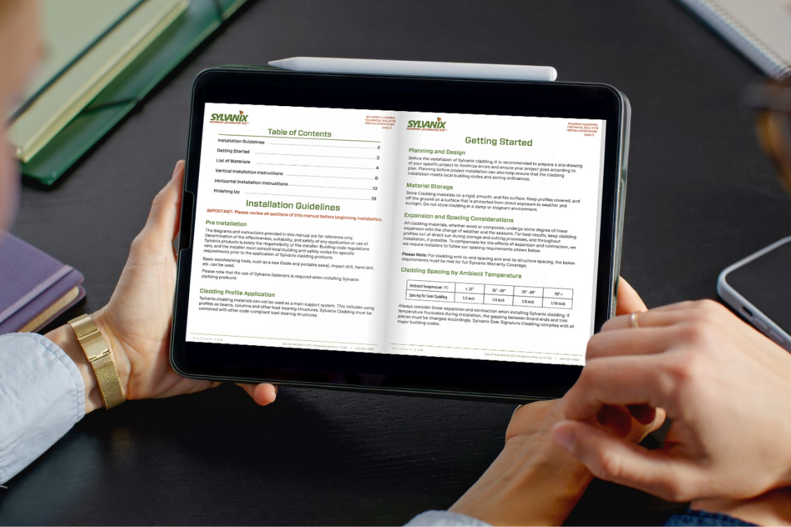 Hands holding a tablet displaying a digital PDF document about Sylwaux installation guidelines, with a closed notebook nearby on a dark table.