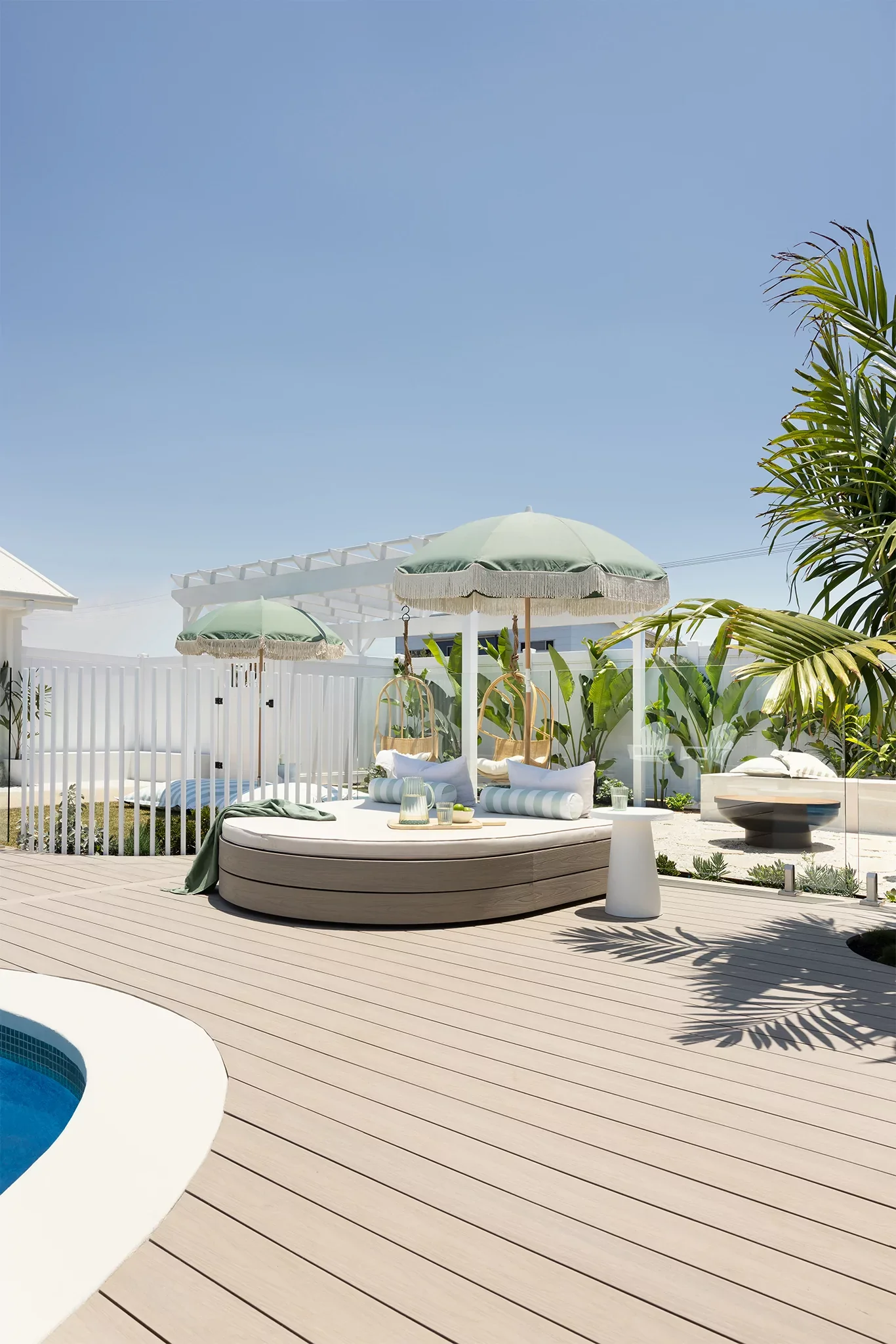 A modern outdoor pool area with a hot tub, umbrellas for shade, white fencing, green plants, and a clear blue sky.