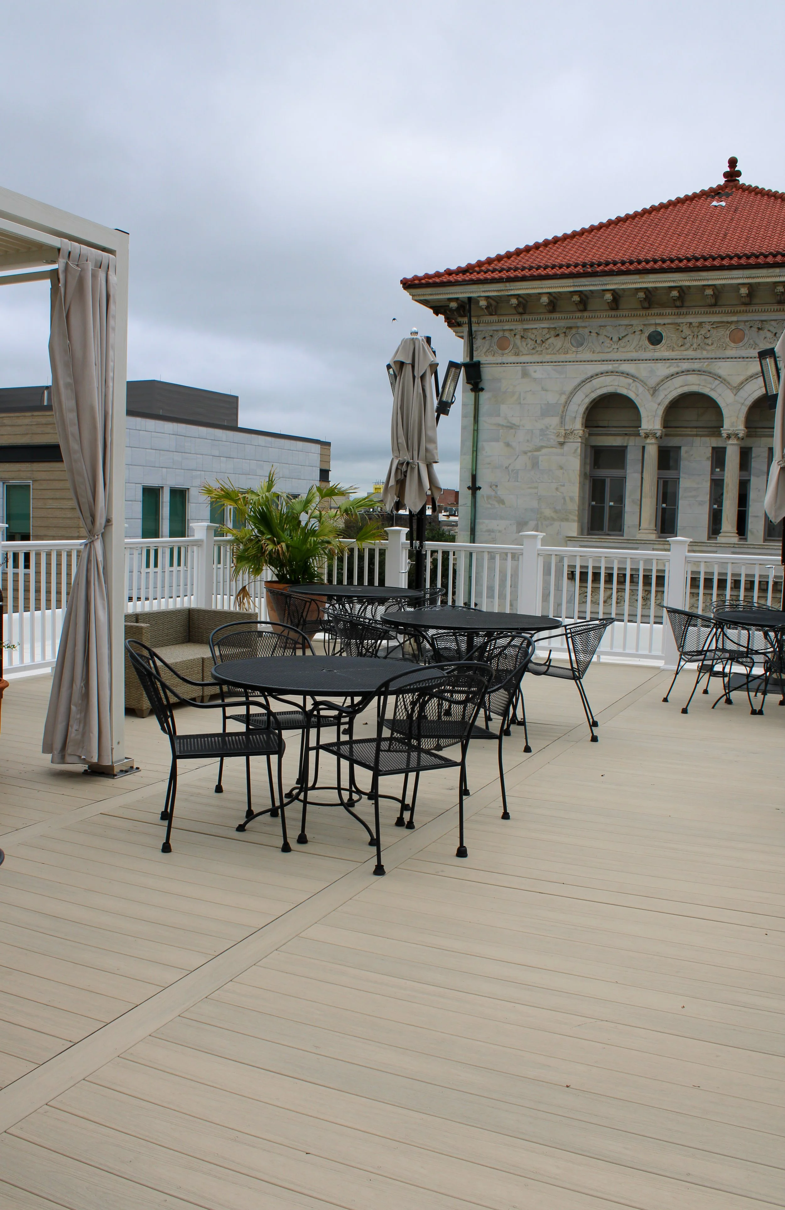 Outdoor patio with black metal tables and chairs, beige wooden flooring, large potted palm plant, beige cabana with curtains, and historic building with red tiled roof and arched windows under a cloudy sky.