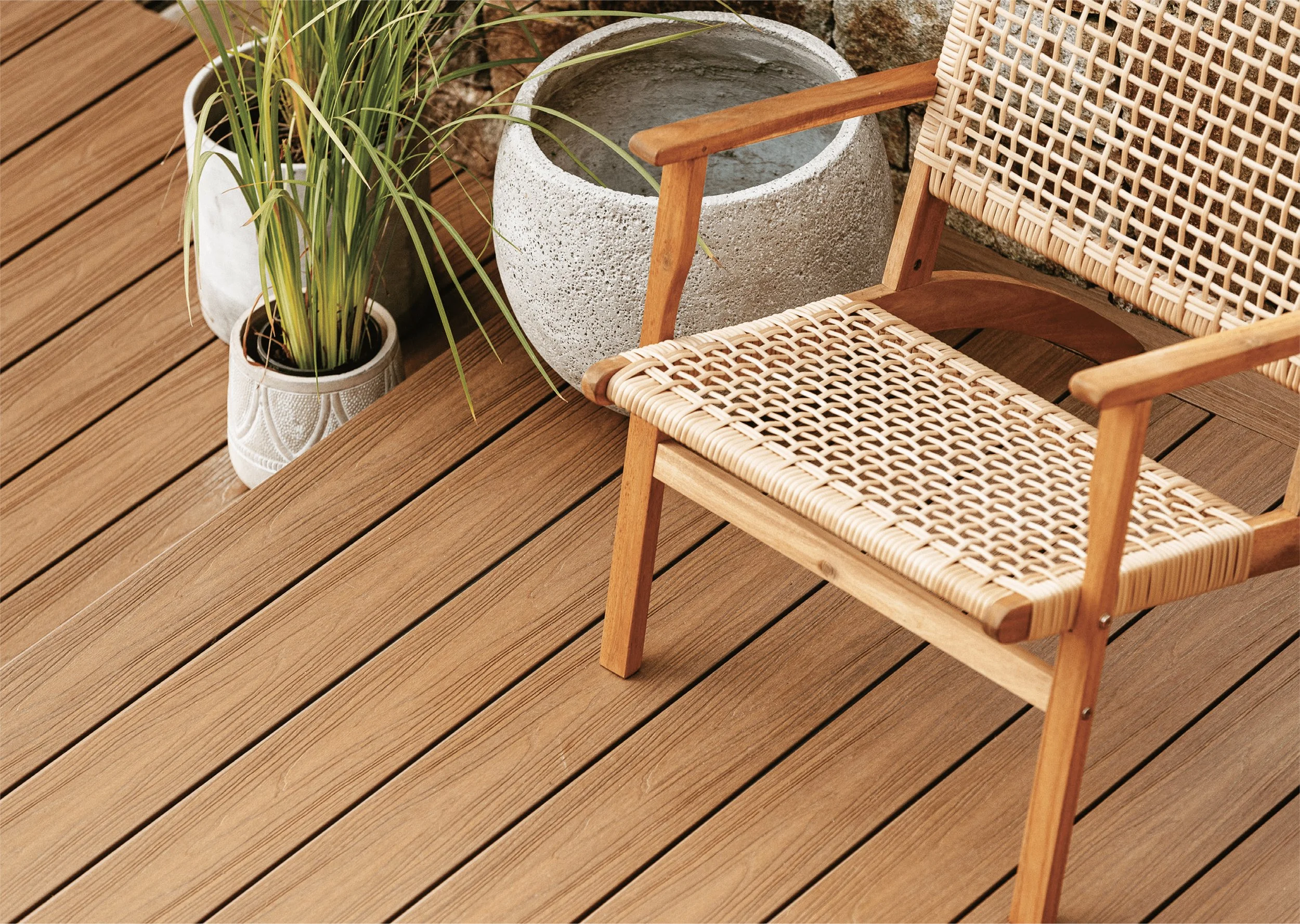 A wooden outdoor deck with a woven cane chair, potted grass, and a large potted concrete planter.