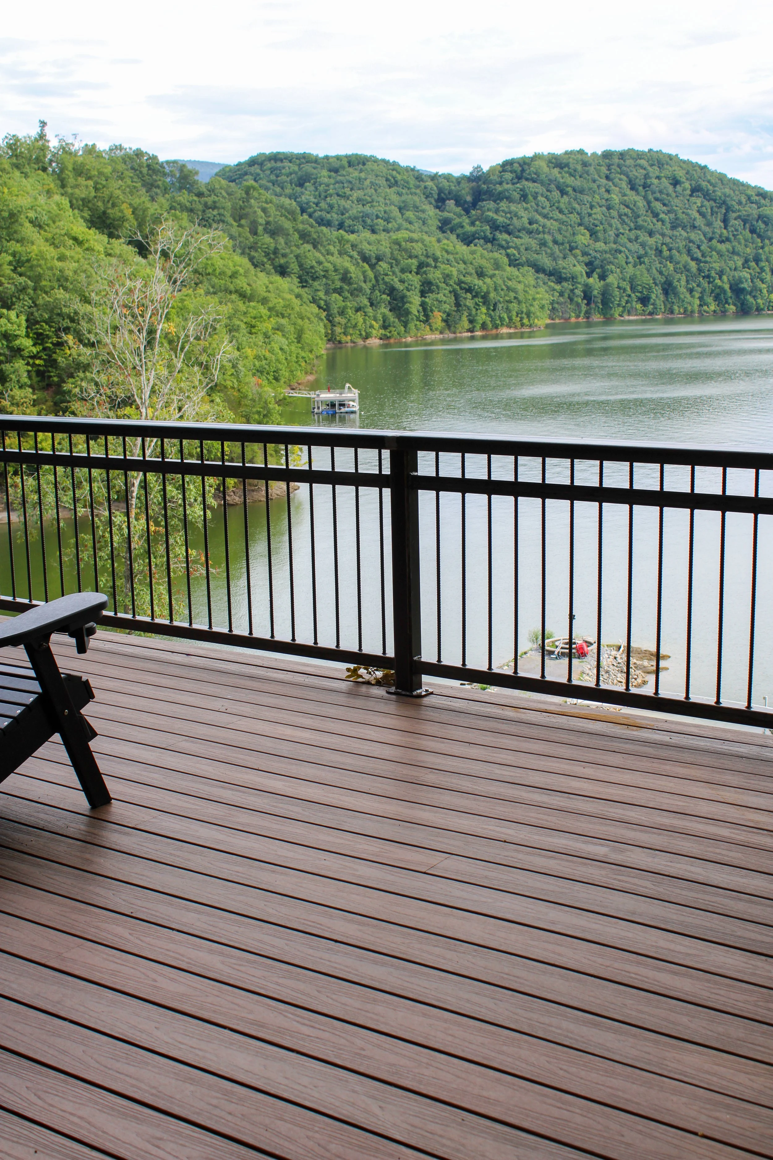A wooden deck with horizontal planks and black railing overlooking a body of water. The landscape includes green trees, hills, and a cloudy sky.