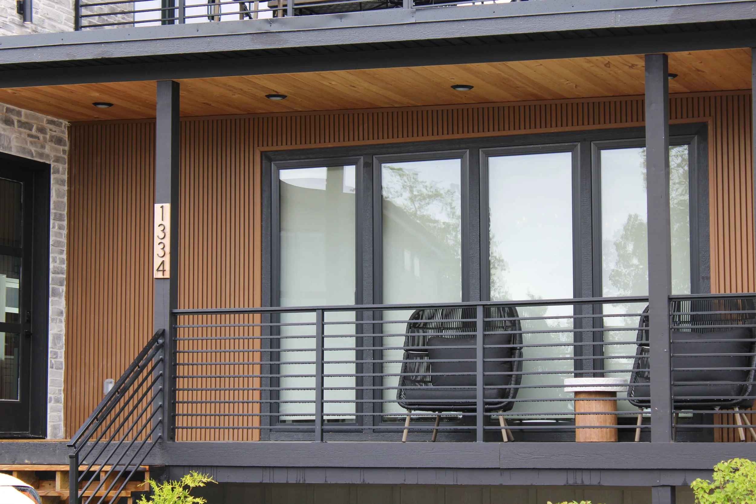 Close-up of a modern apartment balcony with black metal railing, two black chairs, wood-paneled walls, sliding glass doors, and a small section of brick wall.