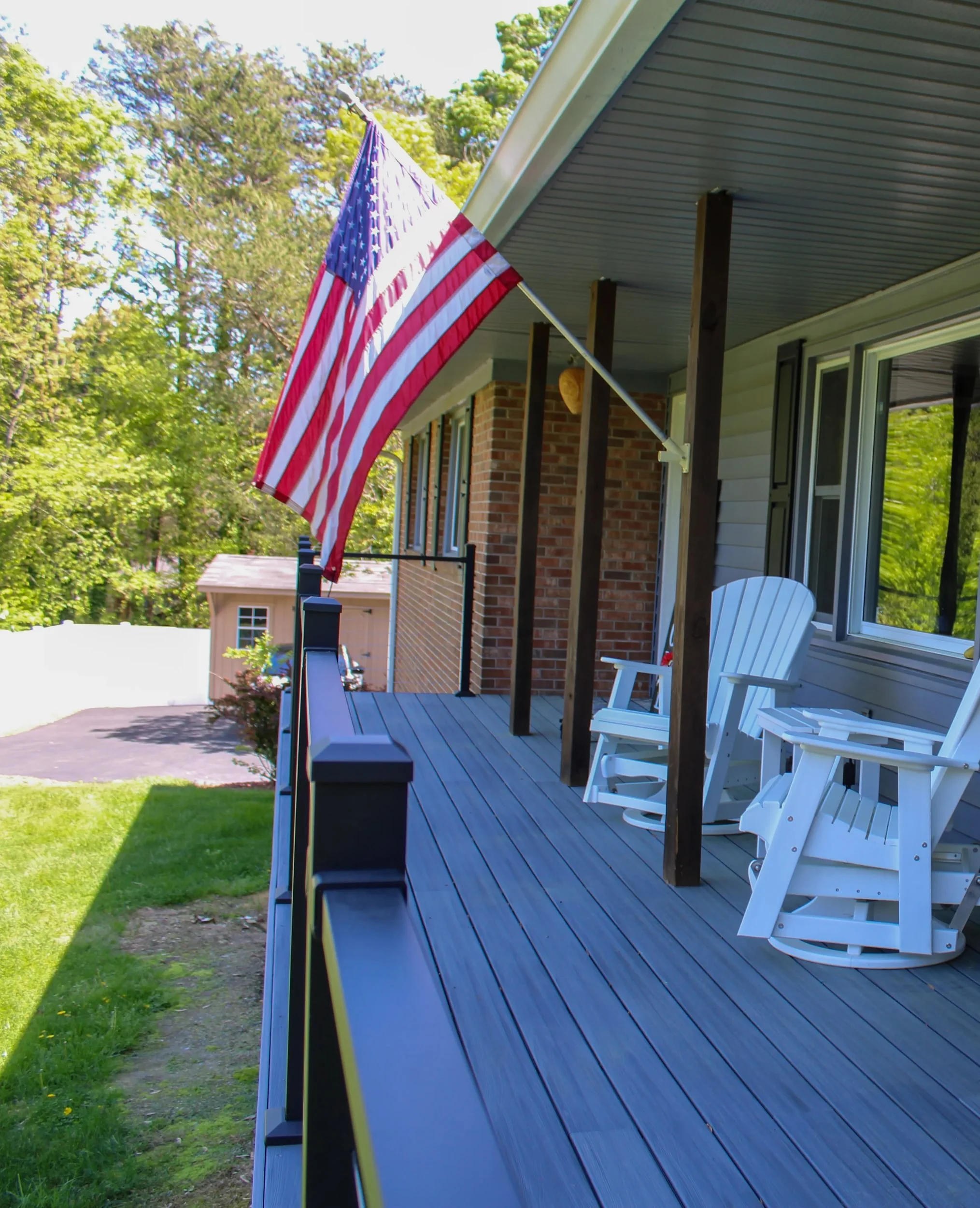 American flag hanging from a porch ceiling with white chairs nearby, green trees in the background, and a blue sky.