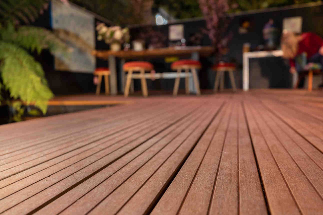 Outdoor wooden deck with some chairs and tables in the background, blurred, and some greenery on the left side.