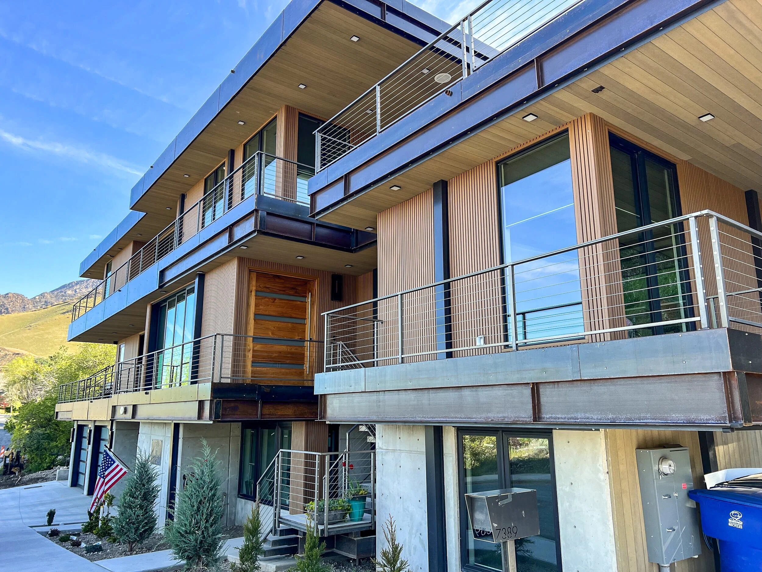 Modern multi-story residential building with balconies, wood paneling, and large glass windows, situated in a scenic area with mountains in the background.