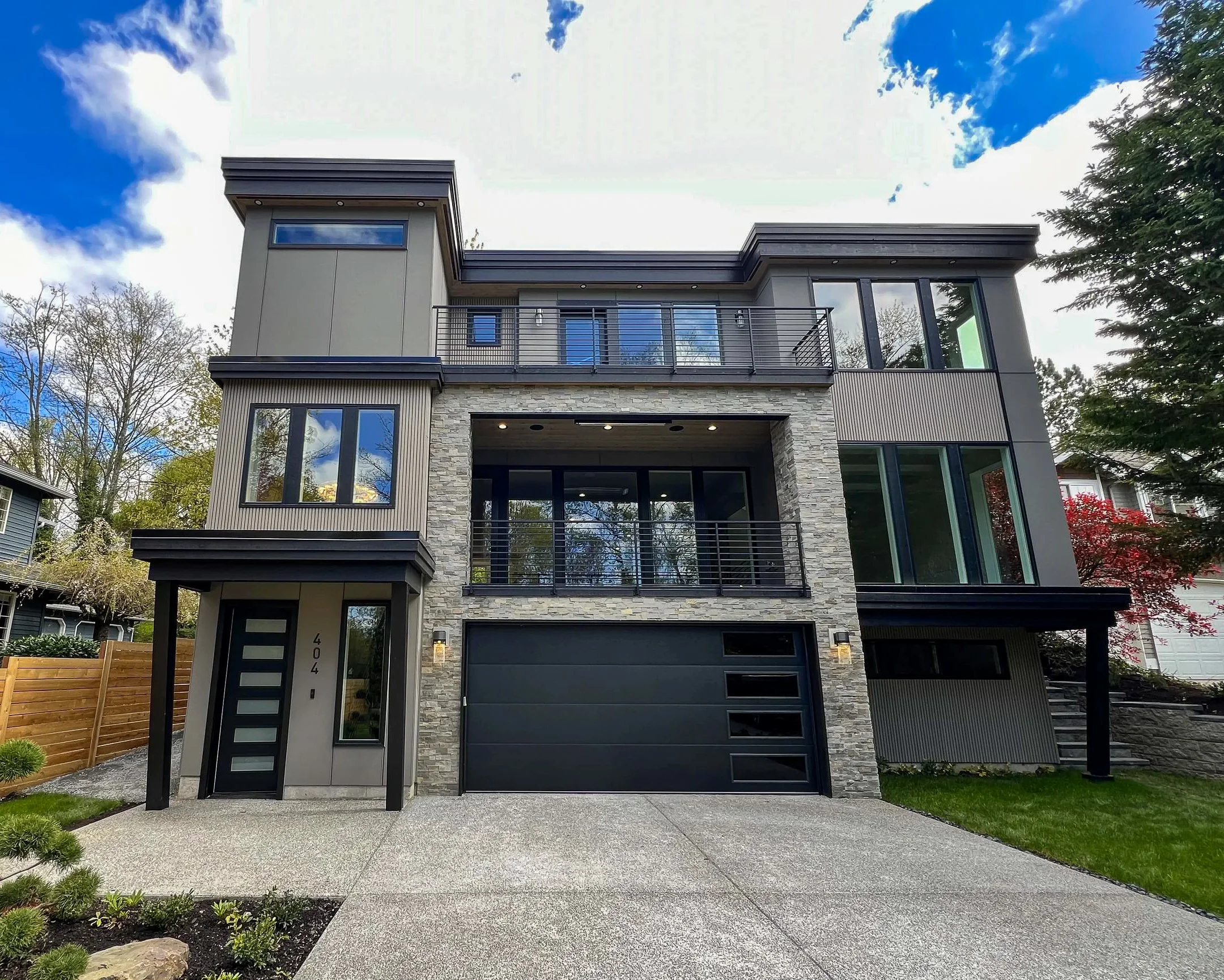 Modern multi-story house with dark garage door, stone and siding exterior, large windows, glass balcony, and landscaped front yard.