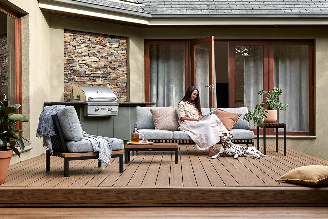 A woman relaxes on an outdoor patio sofa with a Dalmatian dog lying beside her. The patio has wooden flooring, potted plants, a small table with a drink, and is adjacent to a house with large windows and a built-in grill.