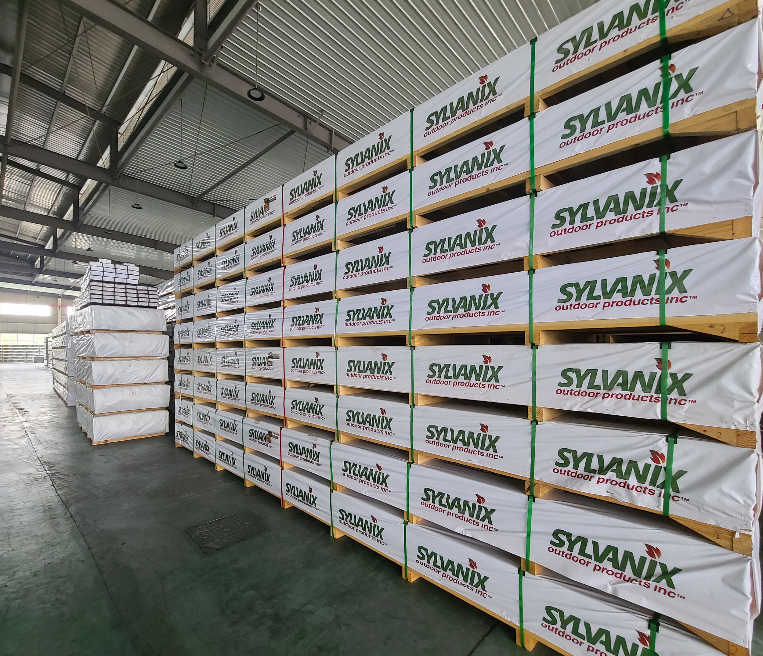 Warehouse with stacks of wood pallets wrapped in white plastic with green and red "SYLVANIX outdoor products Inc" branding.