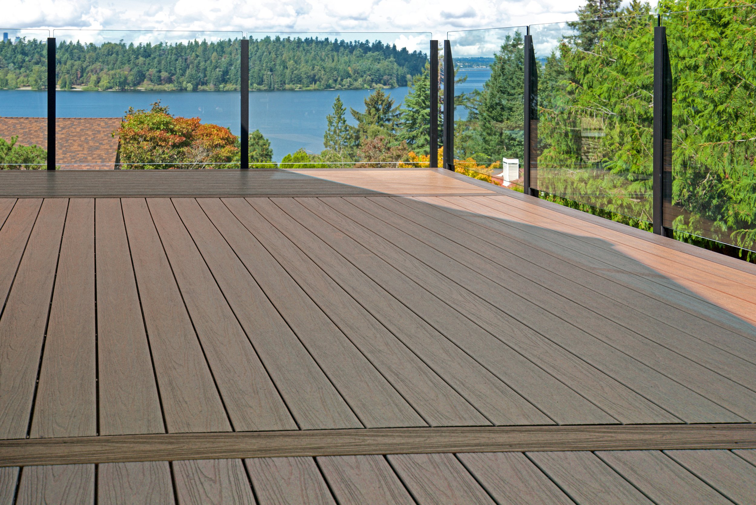 Balcony with wooden flooring and glass railings overlooking a scenic view of a lake, dense trees, and a partly cloudy sky.