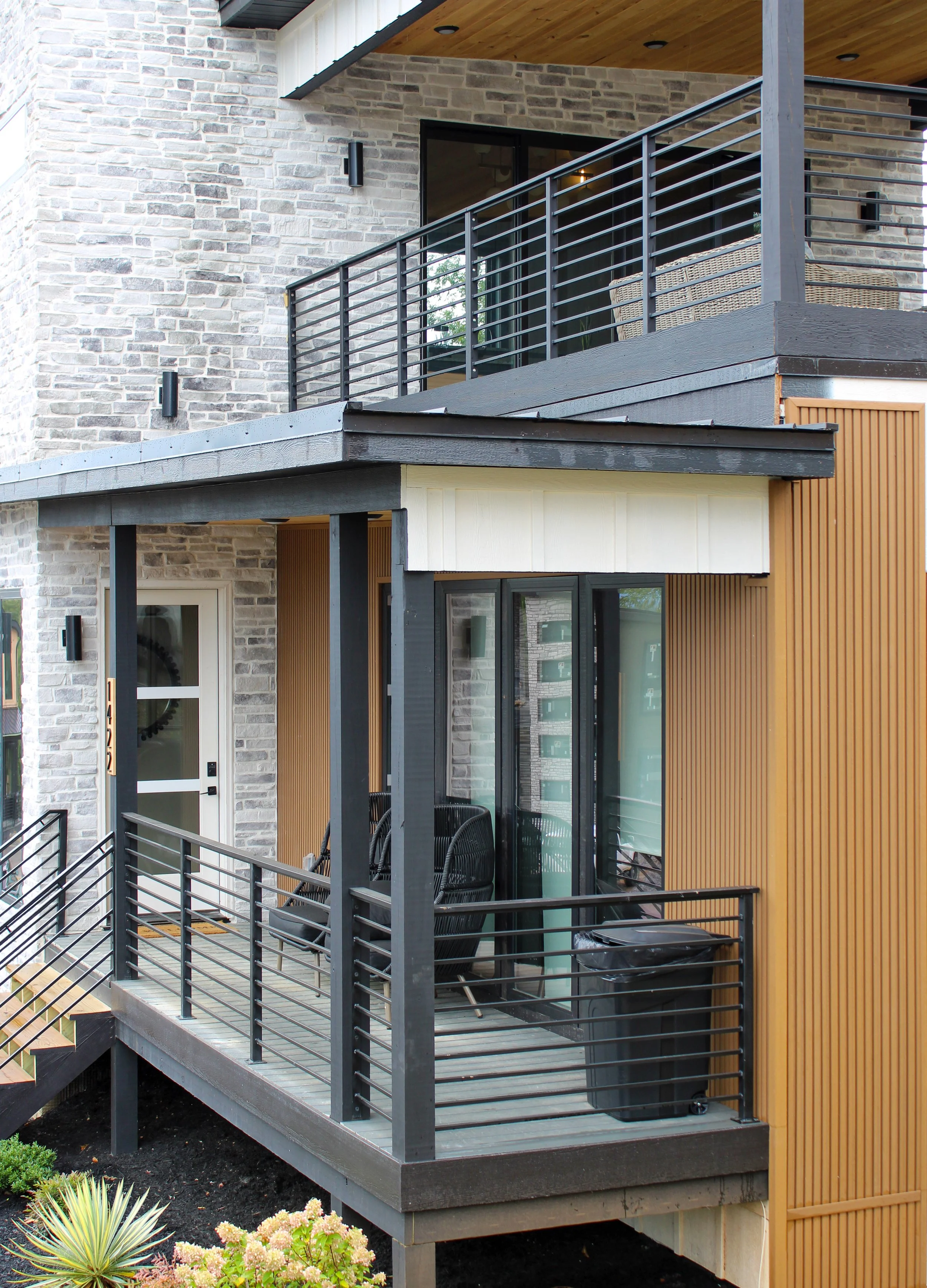 Modern residential balcony with black metal railing, wicker chairs, and sliding glass door, attached to a house with stone and wood exterior, overlooking a garden.