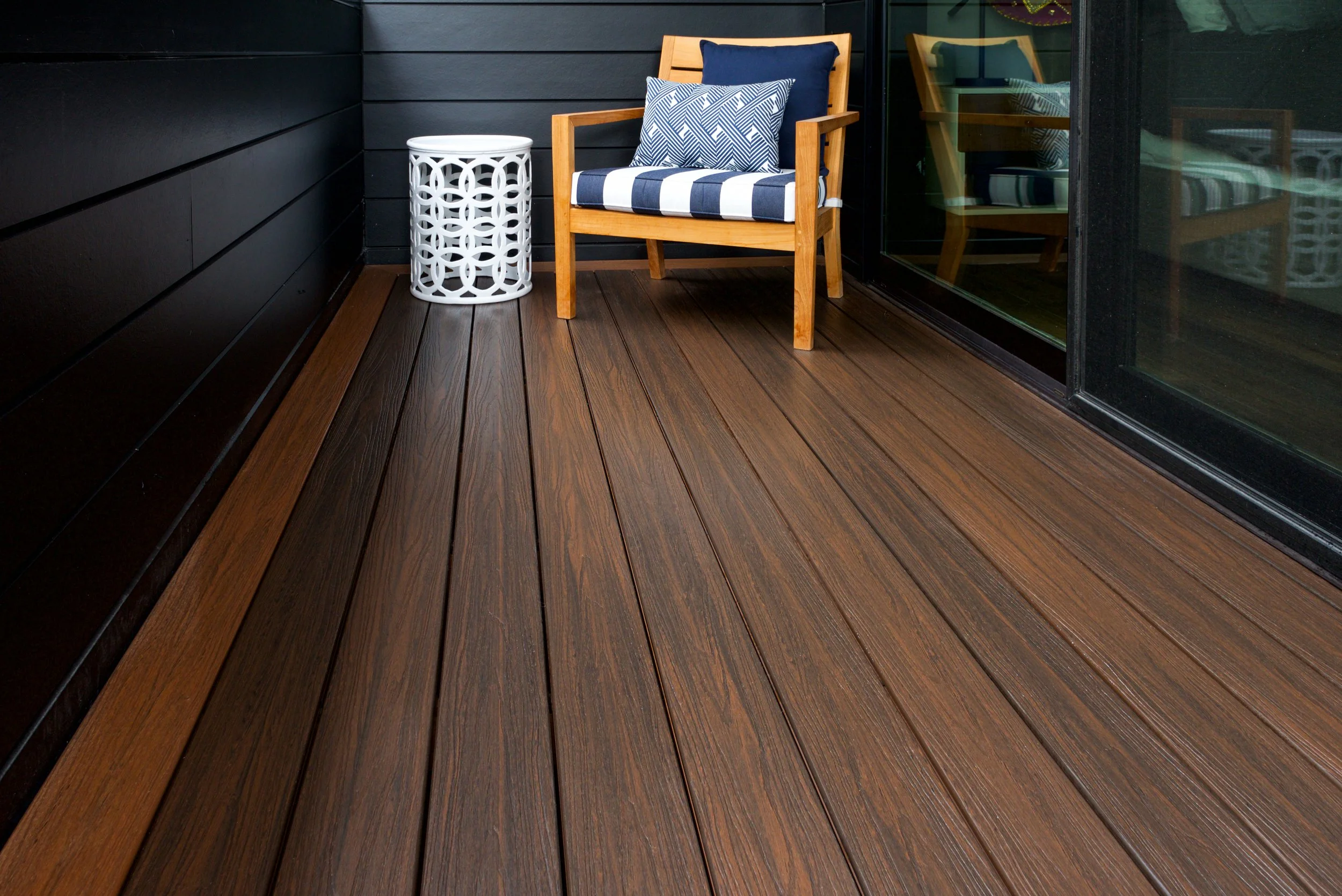 A wooden outdoor chair with blue and white striped cushions and pillows, next to a white lattice side table on a balcony with wood flooring, black wall, and glass sliding door.