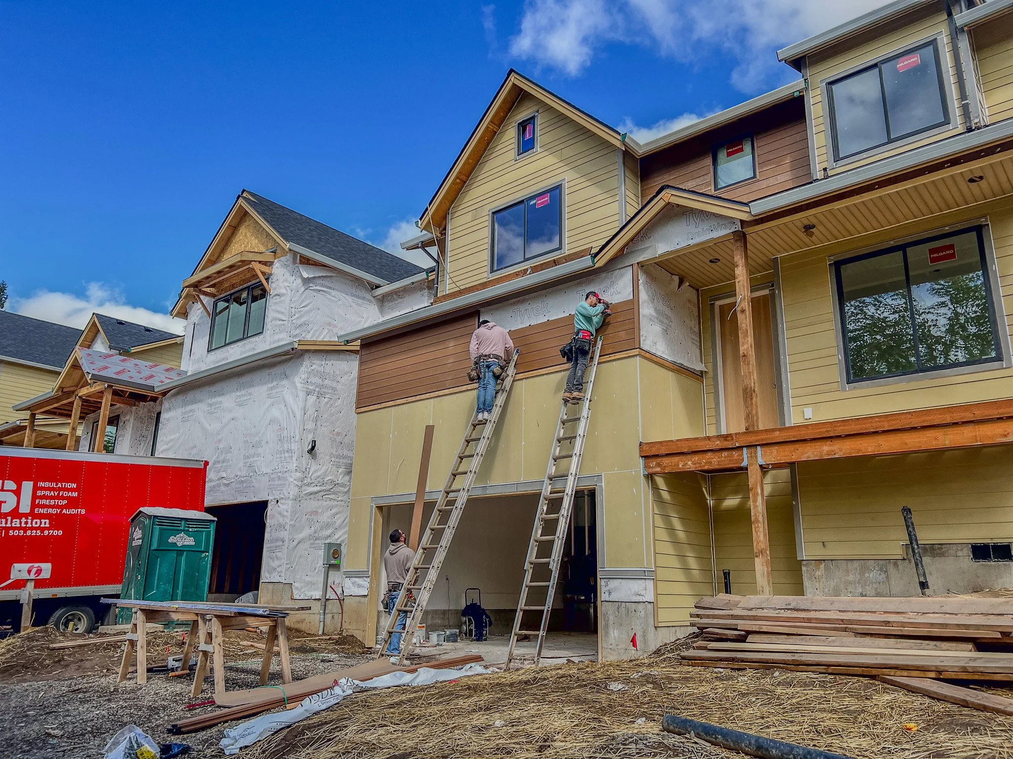 Construction workers building or renovating a multi-story house with exterior siding, windows, and scaffolding. Two workers are on ladders working on the upper part of the house. Building materials and equipment are visible around the site, with a blue sky overhead.
