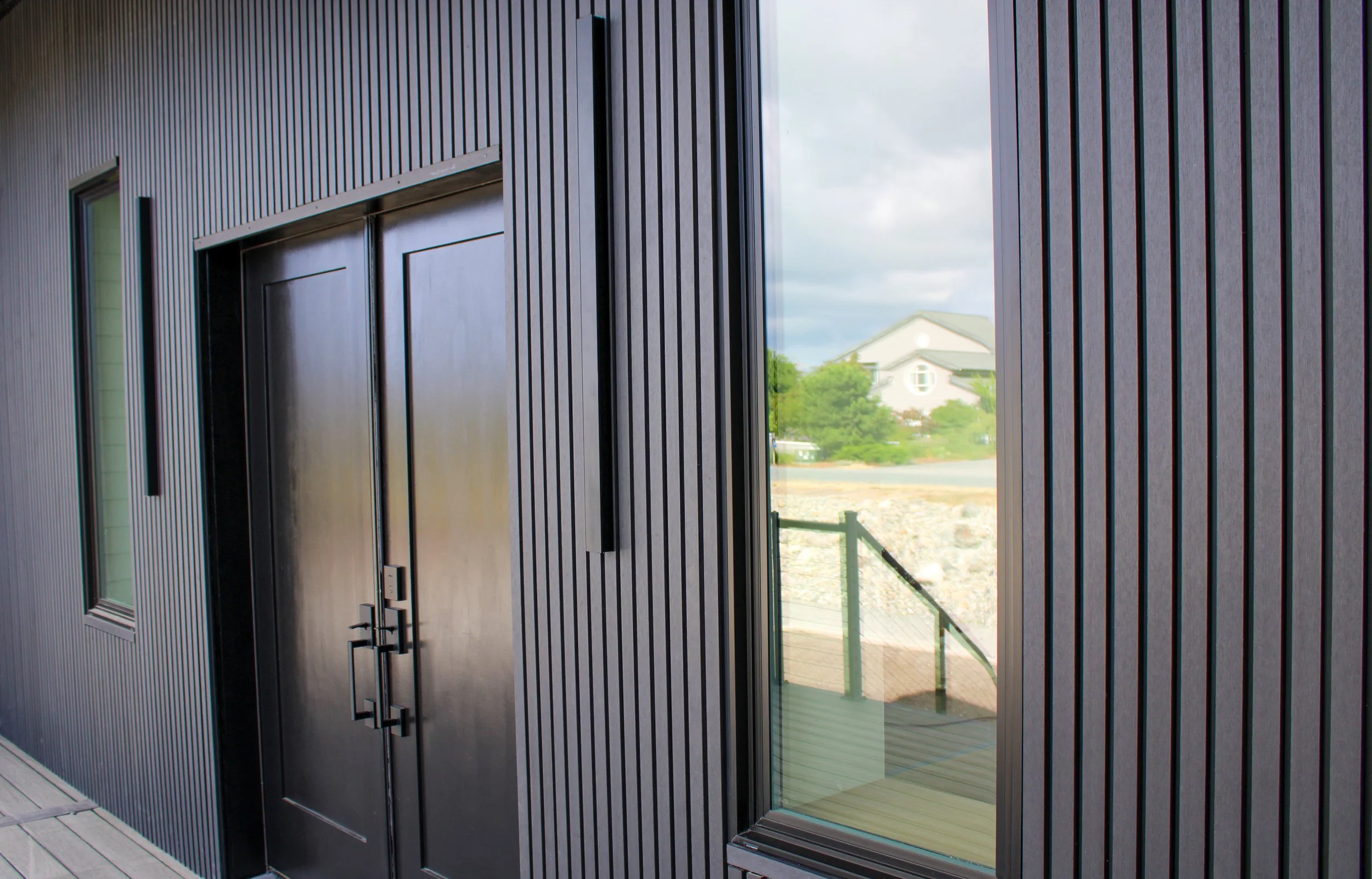 Modern building exterior with vertical black wood siding, large glass windows, and a sliding glass door, overlooking a backyard with green trees and a neighboring house in the distance.