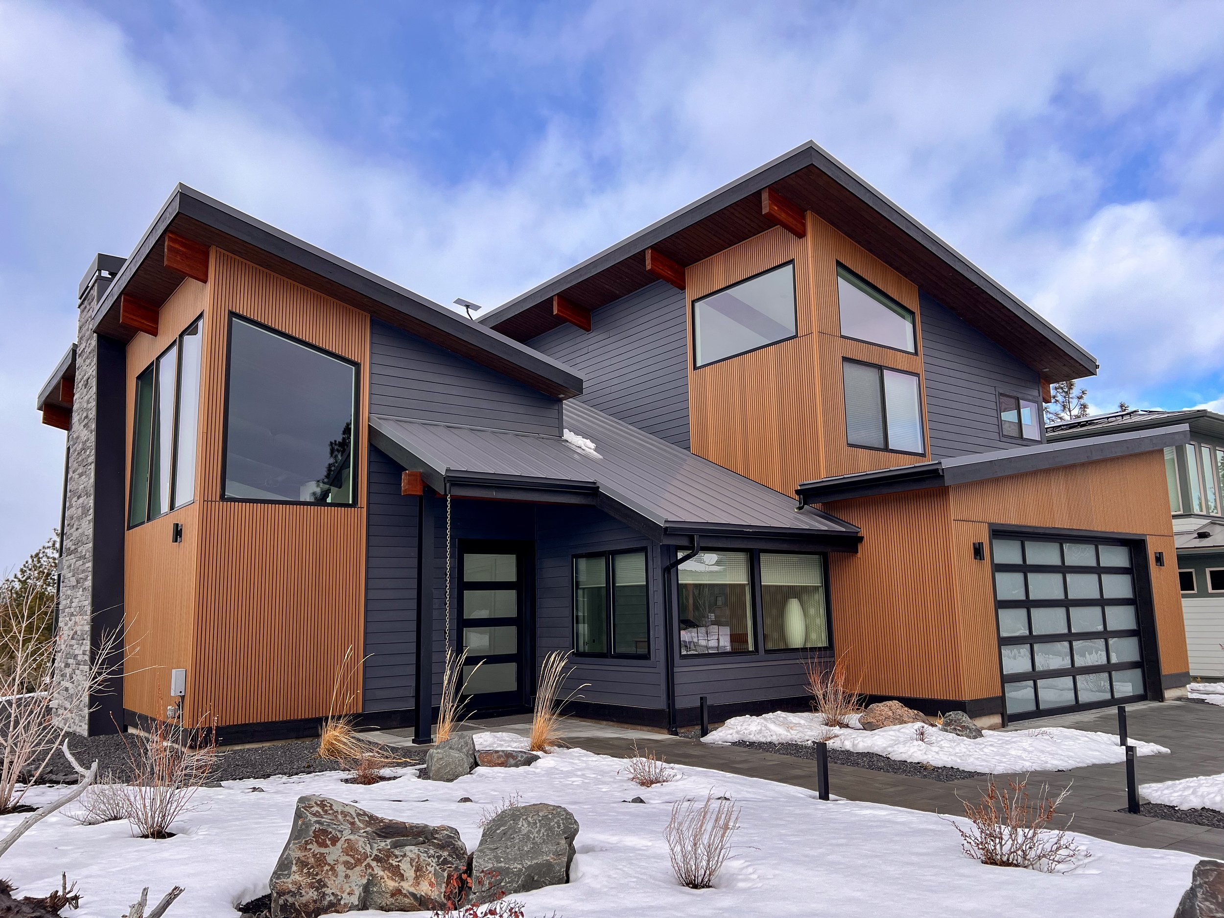 Modern house with wood paneling and large windows, snow on the ground, and a cloudy sky.