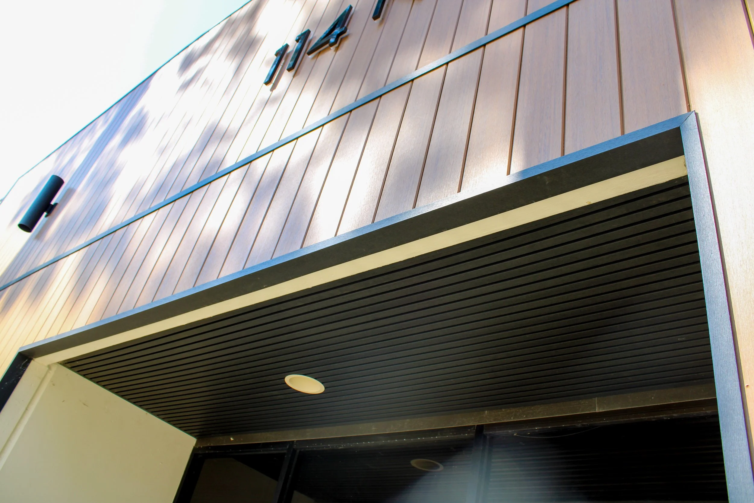 Close-up of a modern building's exterior wall with vertical wooden panels, metal accents, and a black overhang with horizontal slats.